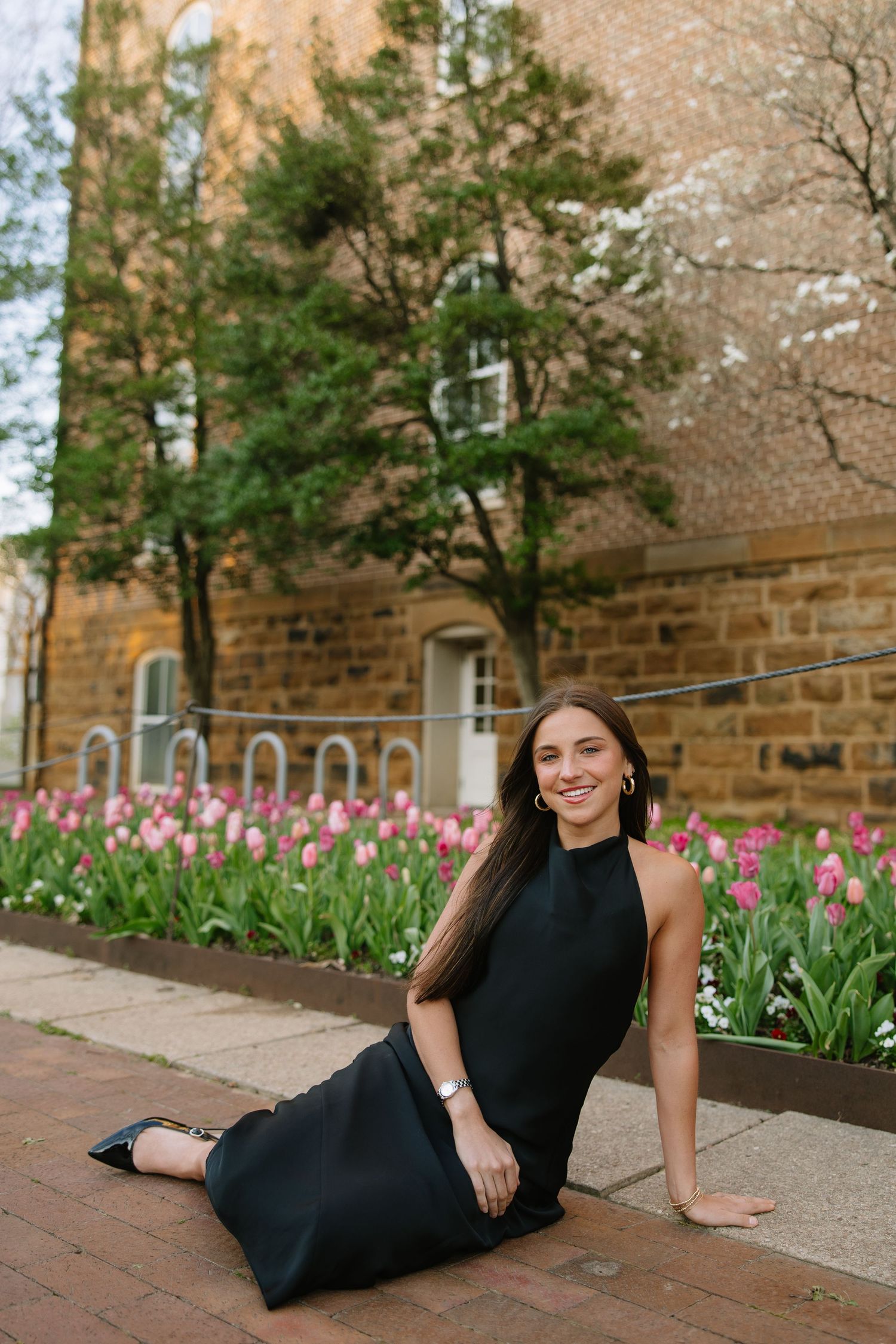 Smiling young woman in black halter dress sitting on brick ledge near pink tulips at university.