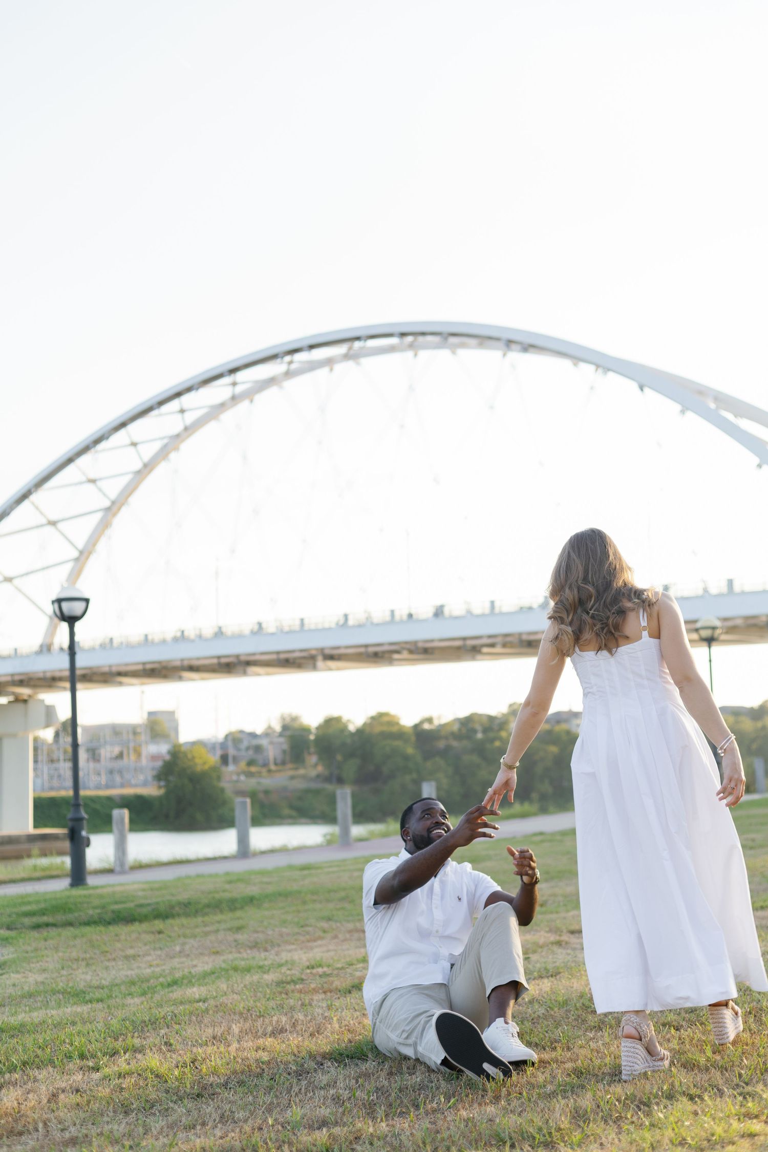 Man sitting on grass reaching up to hold hands with woman in white dress near arched bridge.