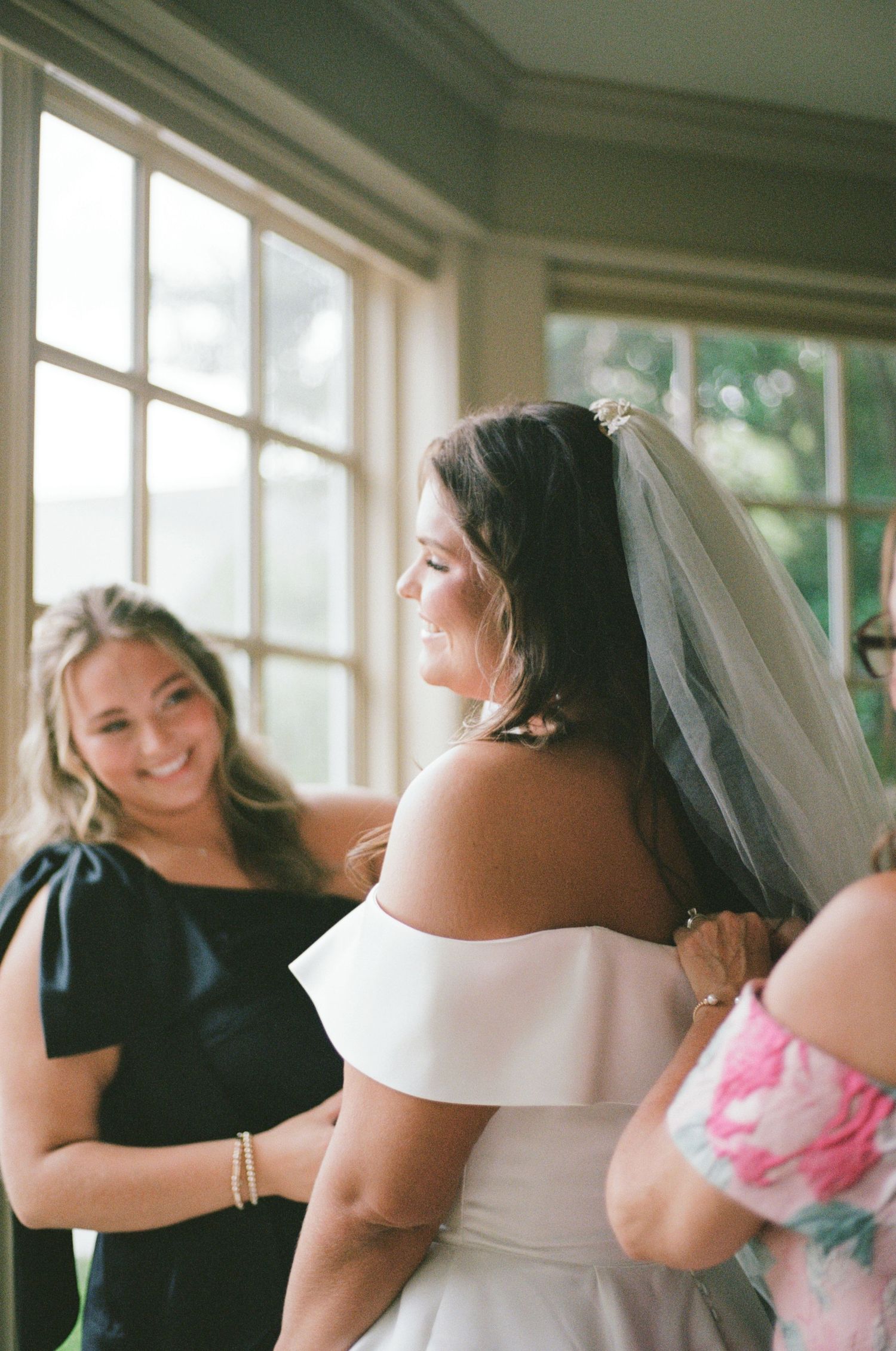 Bride in off-shoulder white gown and veil smiles as bridesmaids help her dress near sunlit windows.