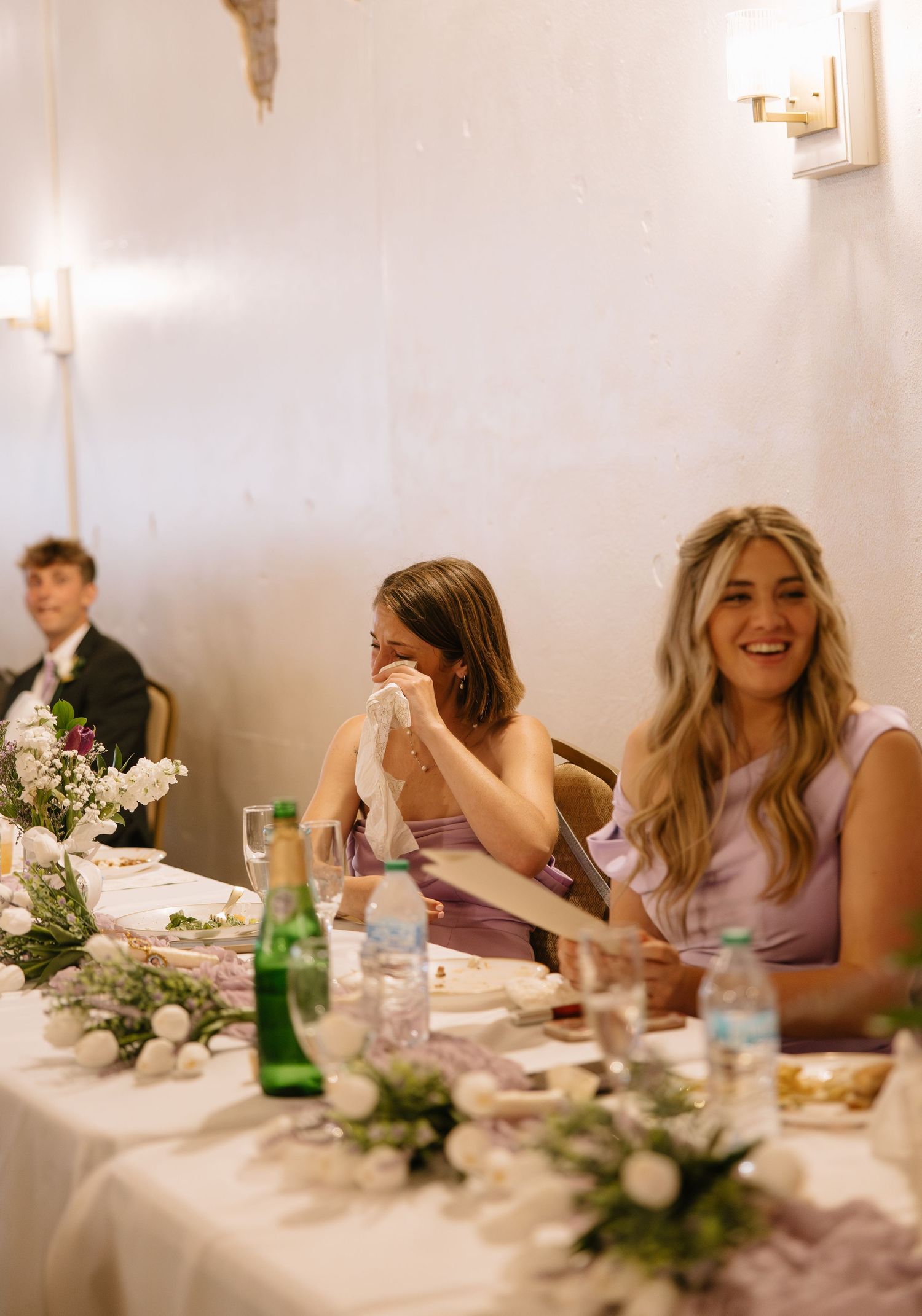 Bridesmaids laughing and crying with joy at a wedding reception table decorated with floral centerpieces.