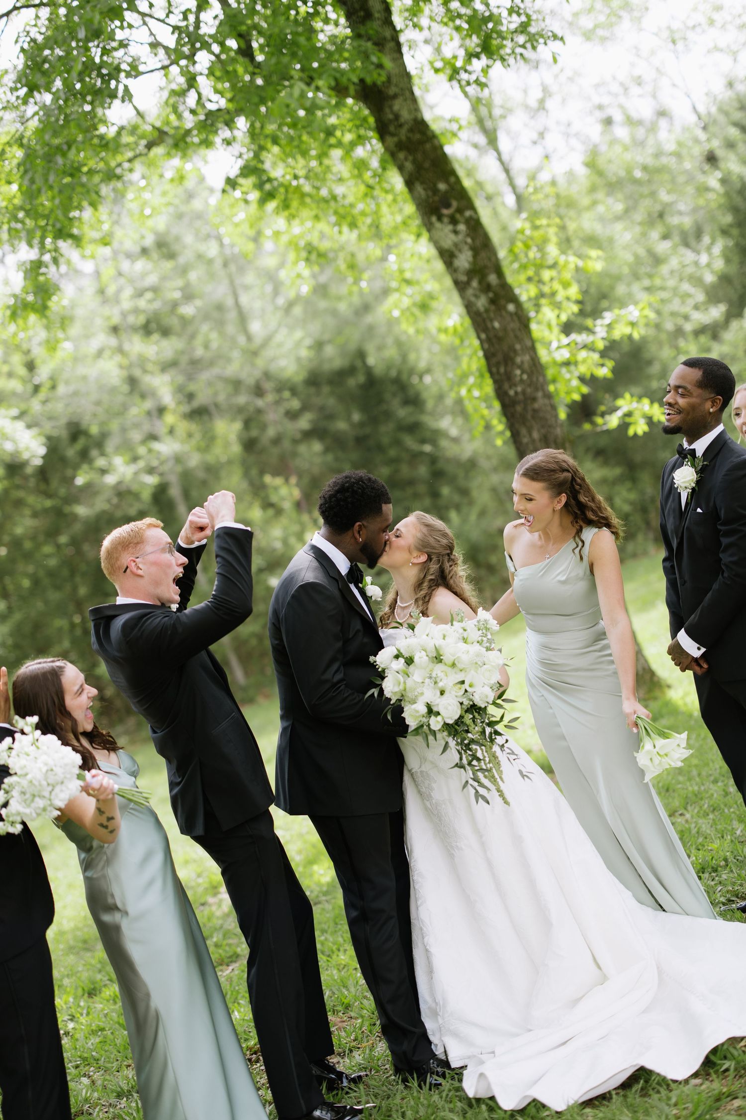 Wedding party cheering as interracial bride and groom share a kiss under a tree in a green outdoor setting.