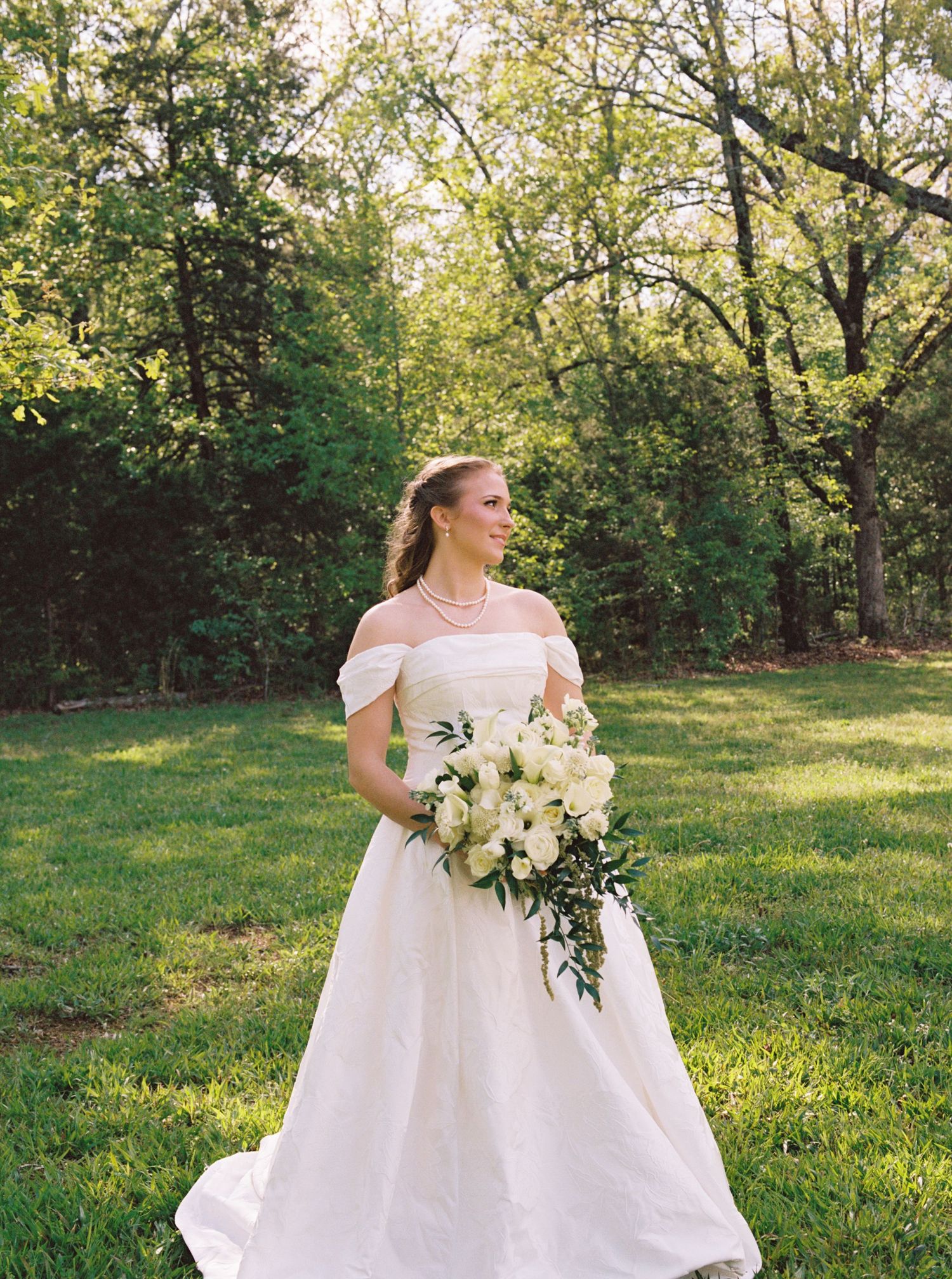 Bride in off-shoulder white gown holding cascading white bouquet in lush green garden.