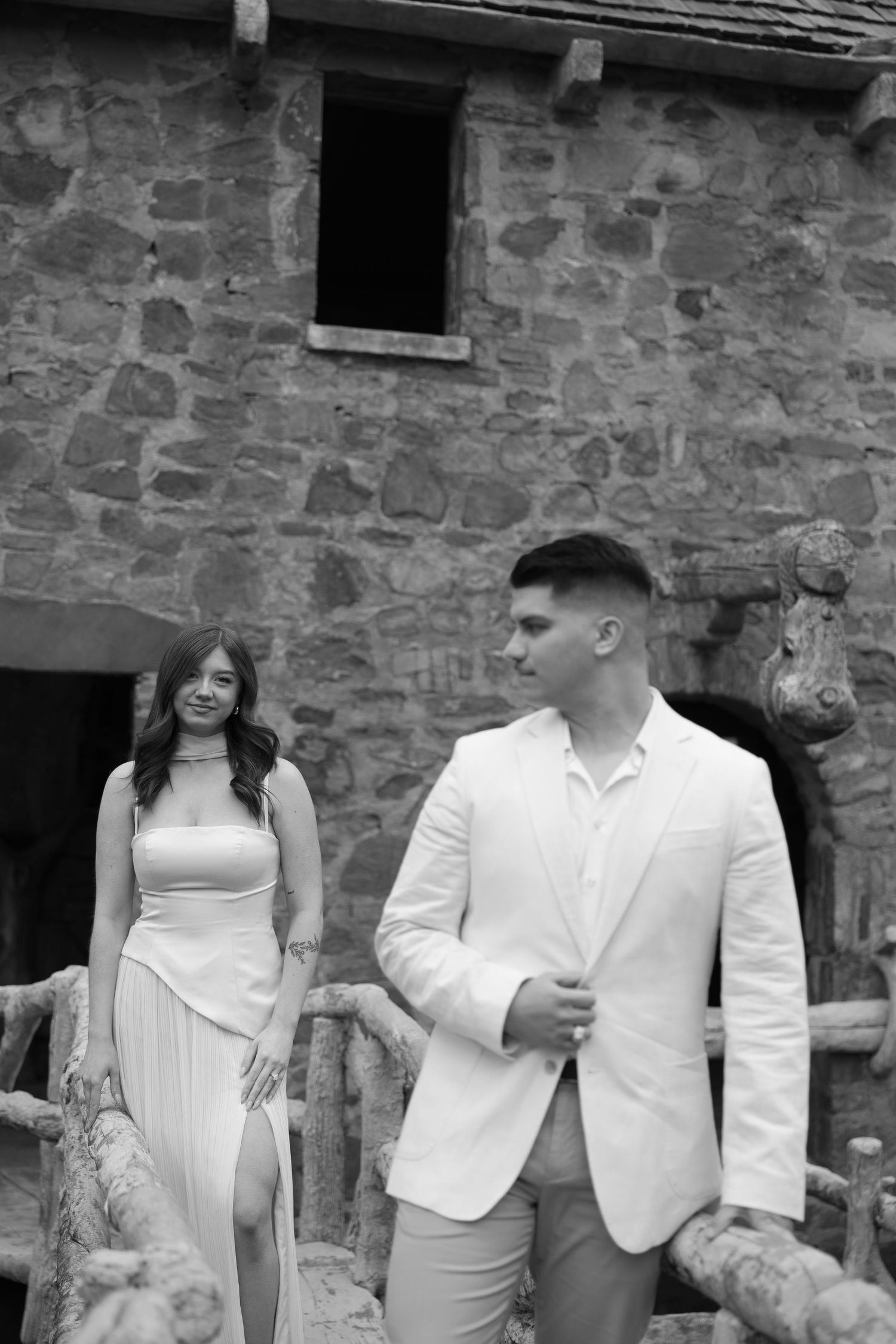 Black and white photo of smiling couple in elegant white attire walking near a rustic stone building.