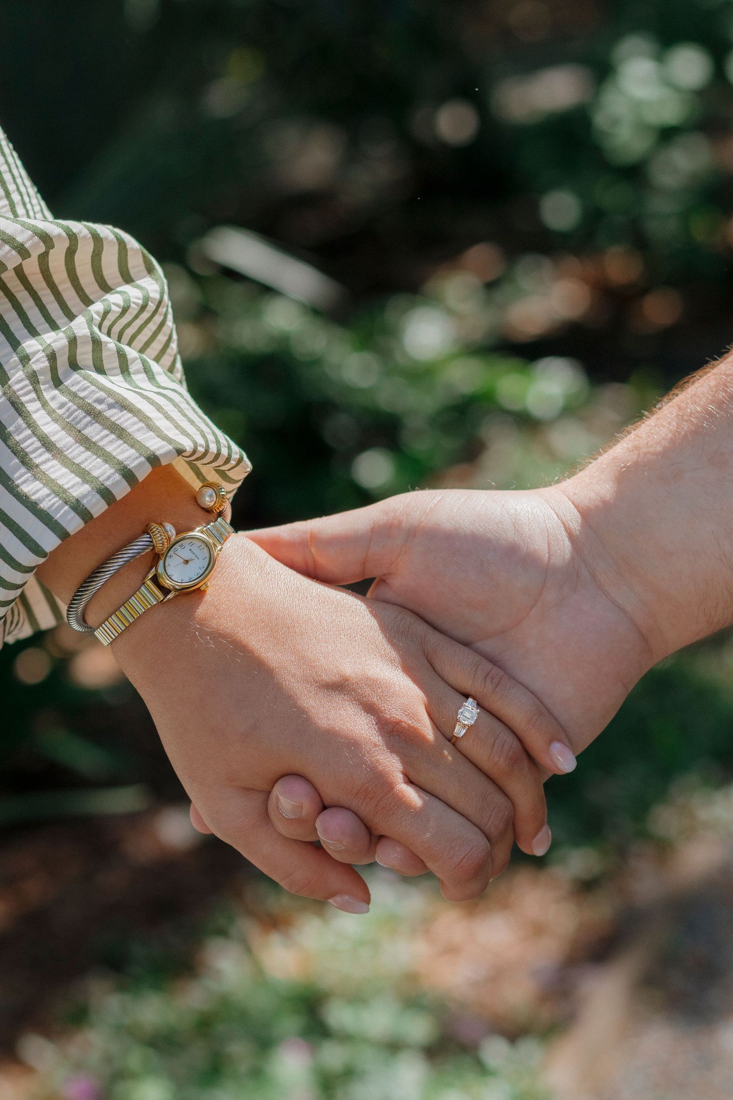 Couple holding hands outdoors, woman wearing a gold watch and striped sleeve, man with a diamond engagement ring.