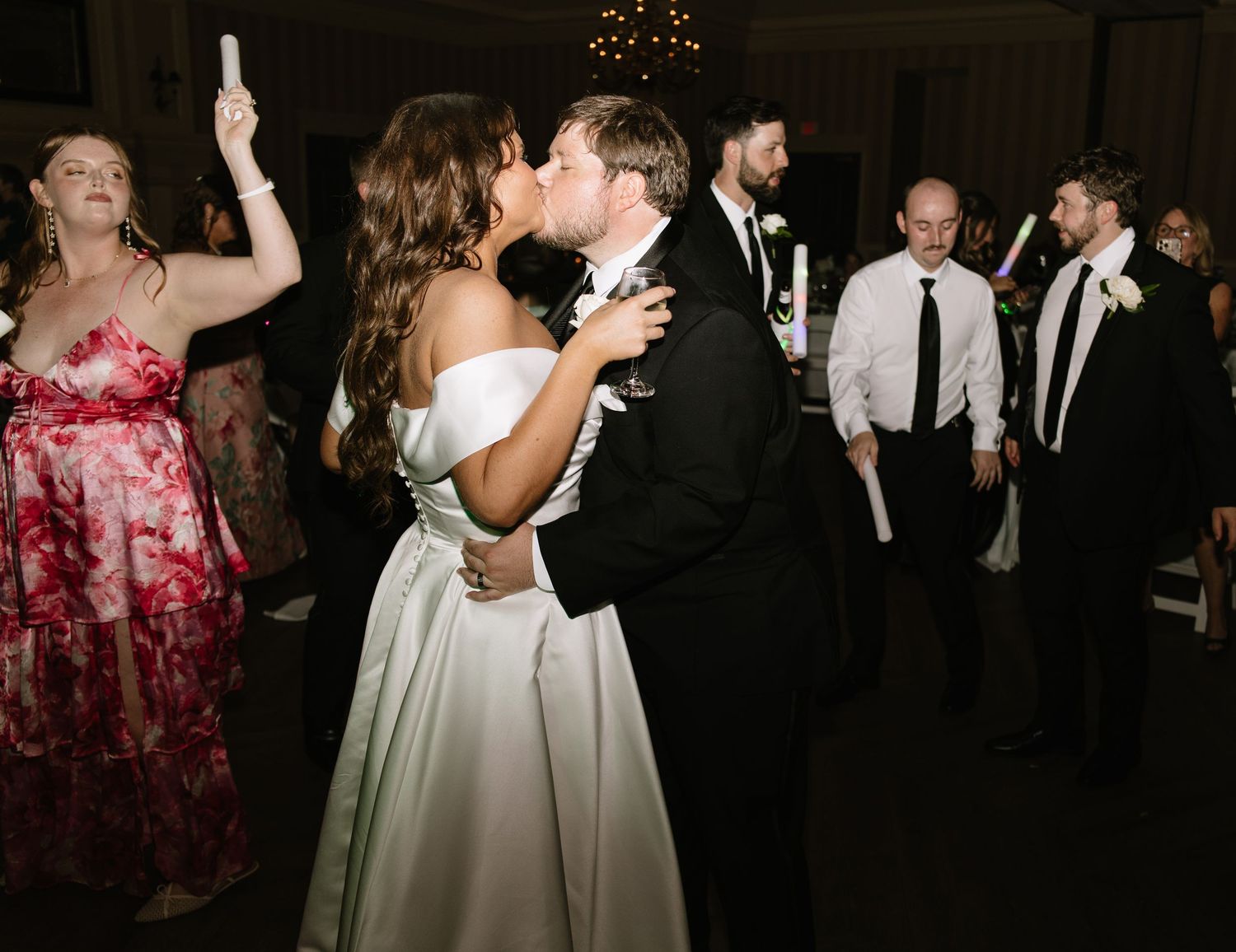 Bride and groom share a kiss on the wedding reception dance floor surrounded by guests in formal attire.
