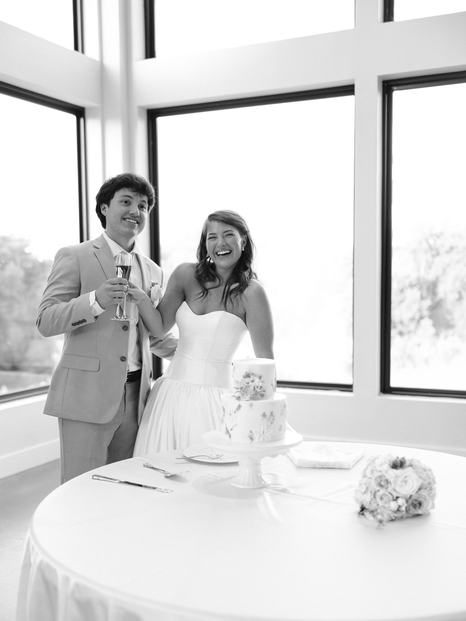 Black and white photo of a smiling bride and groom standing beside their wedding cake during reception toast.