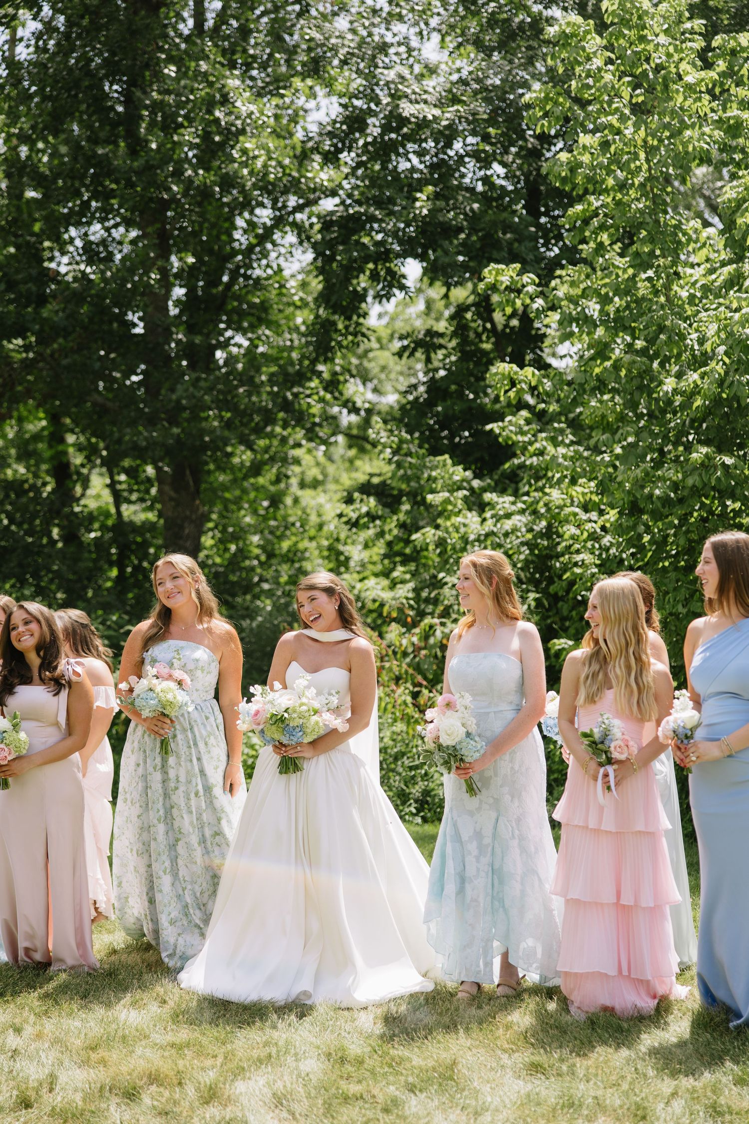 Bride in white gown stands with bridesmaids in pastel dresses holding bouquets outdoors on sunny day.