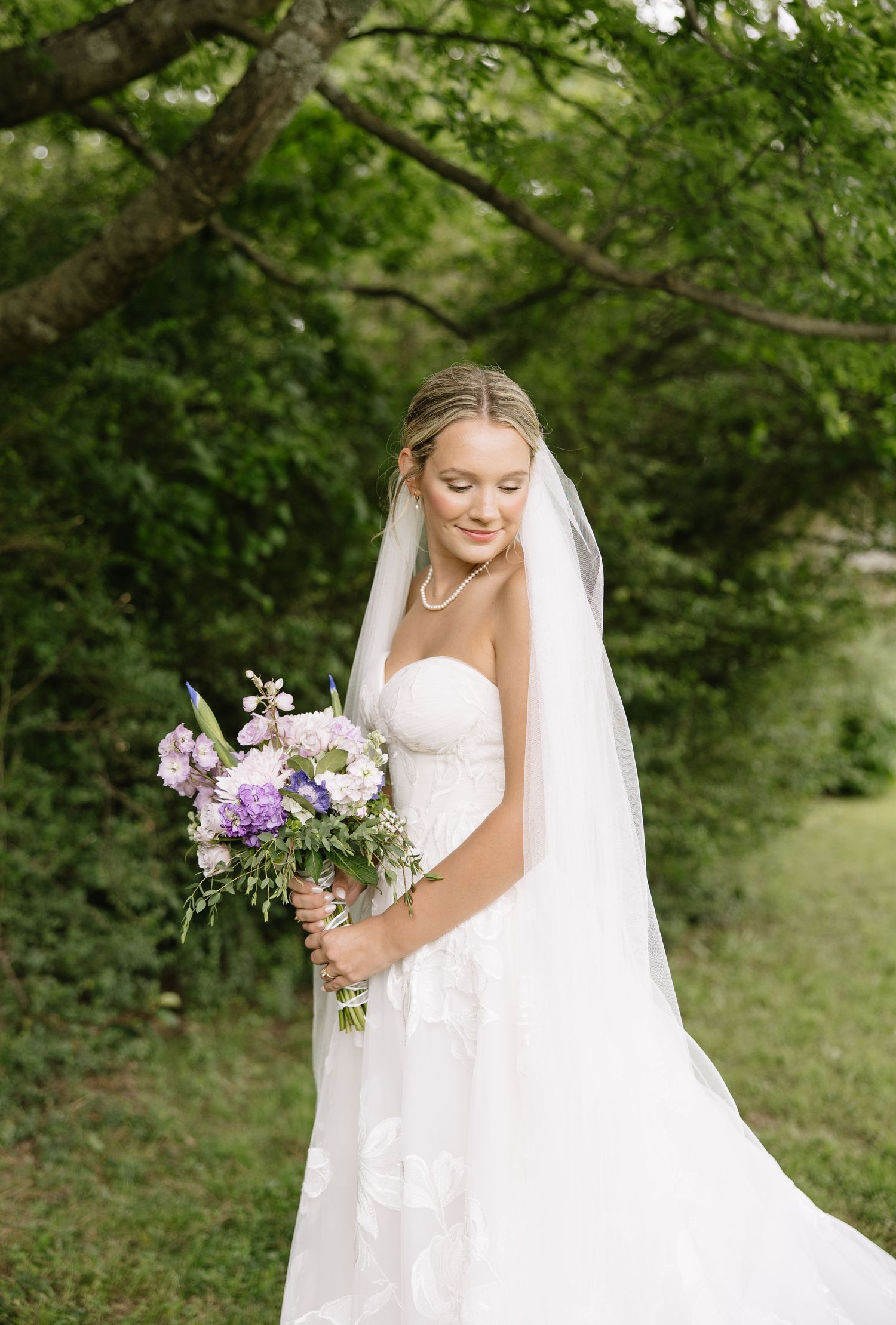 Bride in strapless lace gown and veil holds purple floral bouquet outdoors among lush green trees.