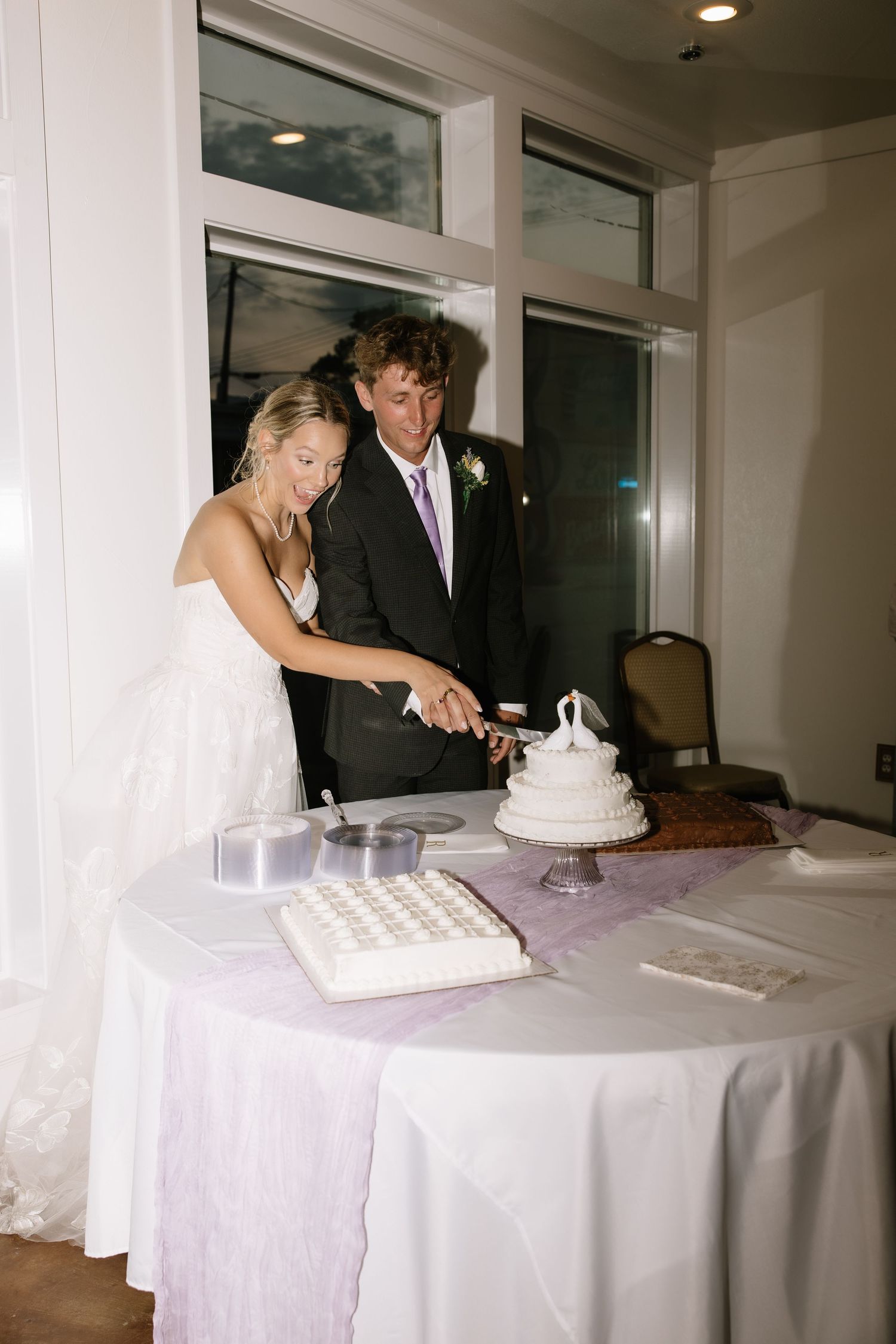 Bride and groom cut white wedding cake together at elegant indoor wedding reception.