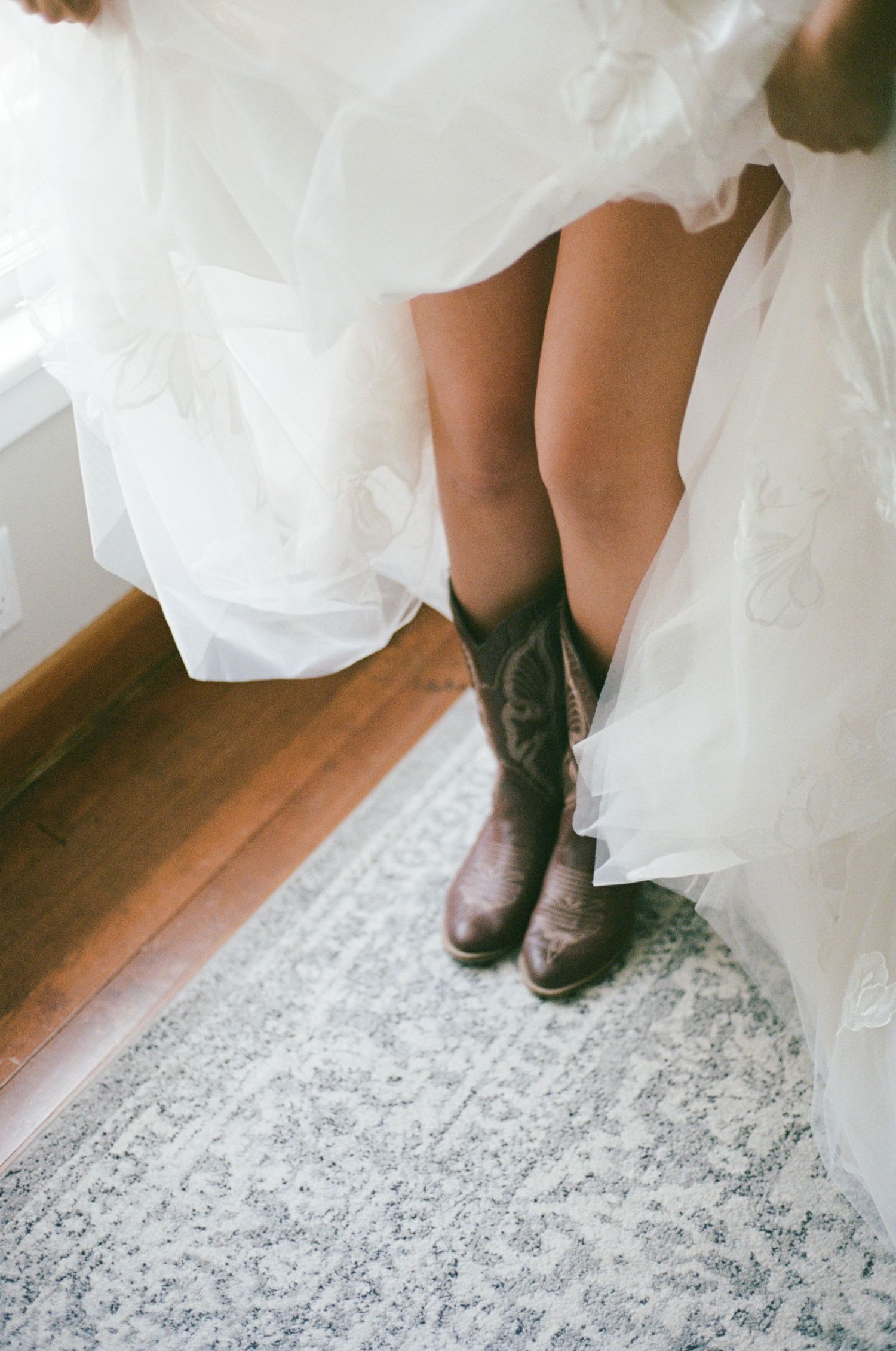 Bride wearing brown cowboy boots under a white tulle wedding dress, standing on a vintage patterned rug.