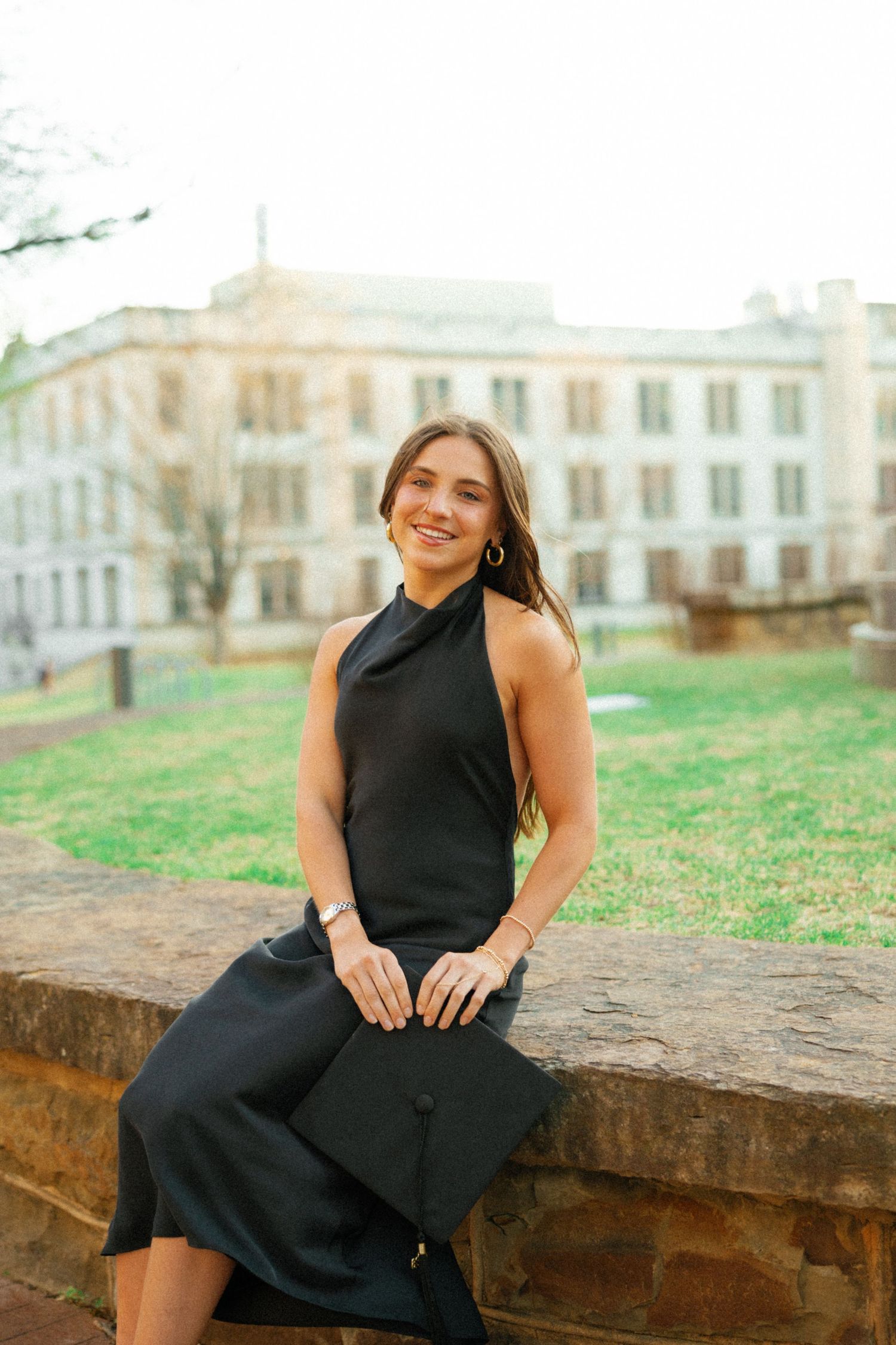 Smiling graduate in black halter dress holding cap while seated on stone wall with campus building behind.