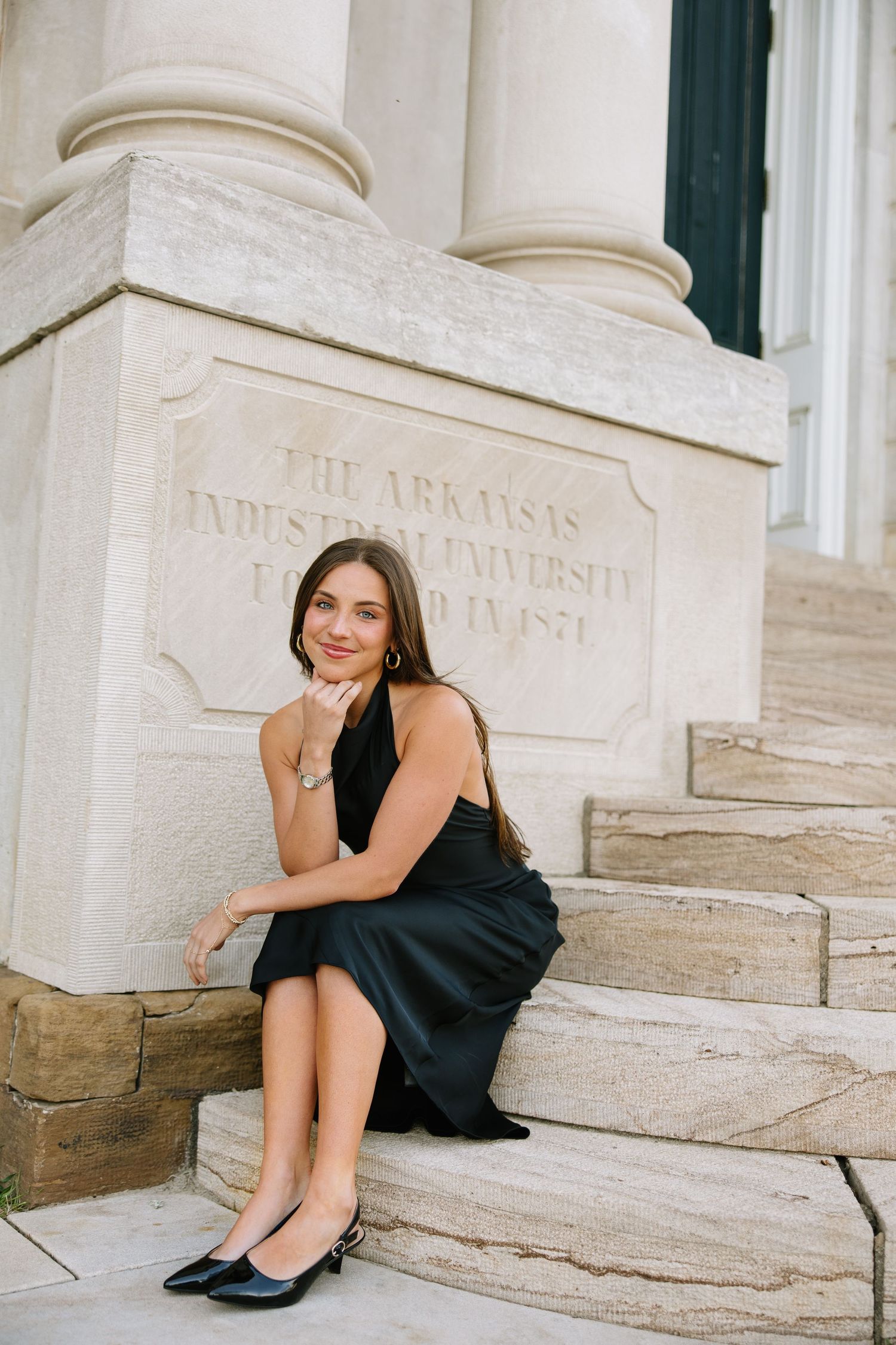 Young woman in black dress sitting on steps beside Arkansas Industrial University founded 1871 inscription.