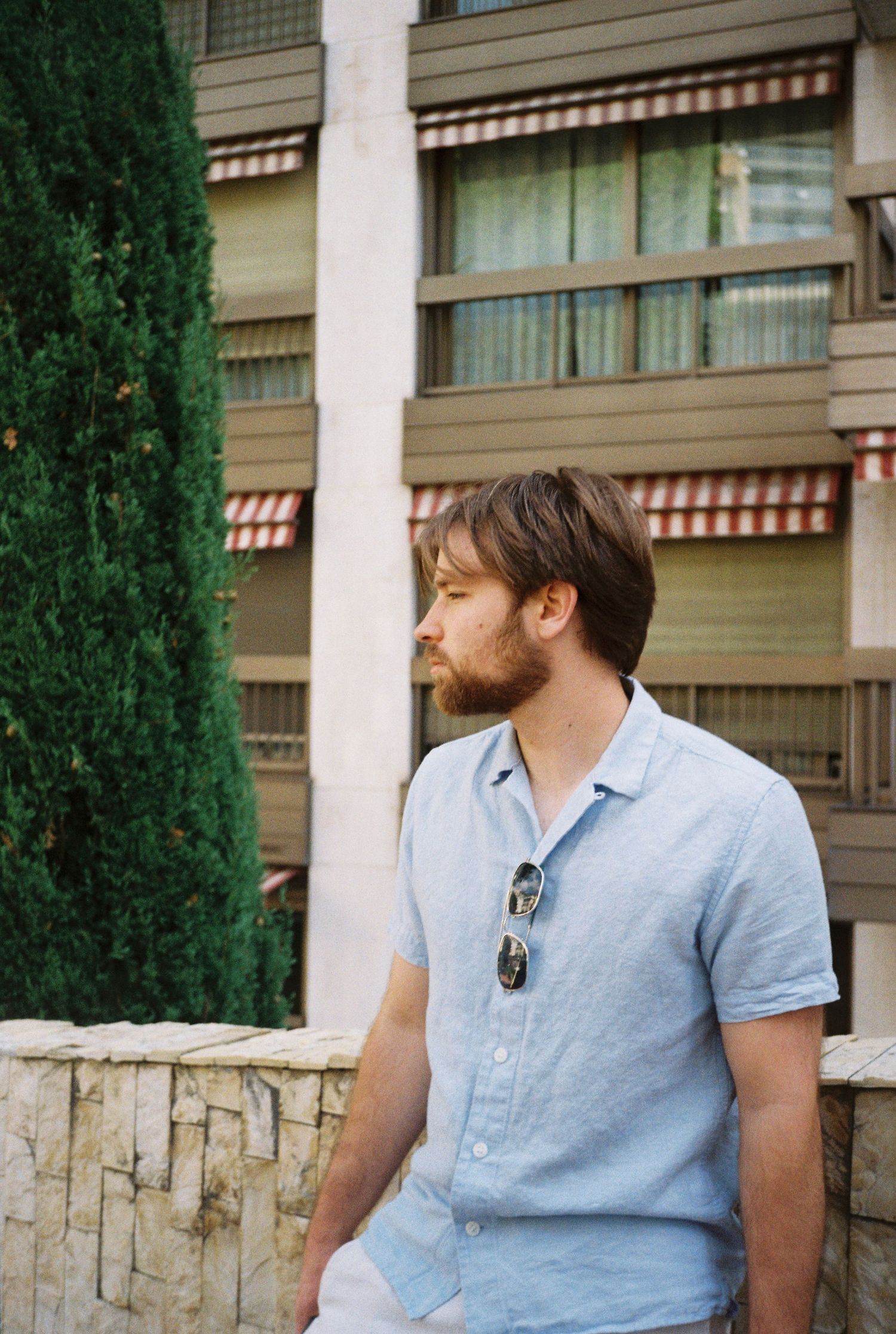 Bearded man in light blue linen shirt sitting outdoors by building with cypress tree.