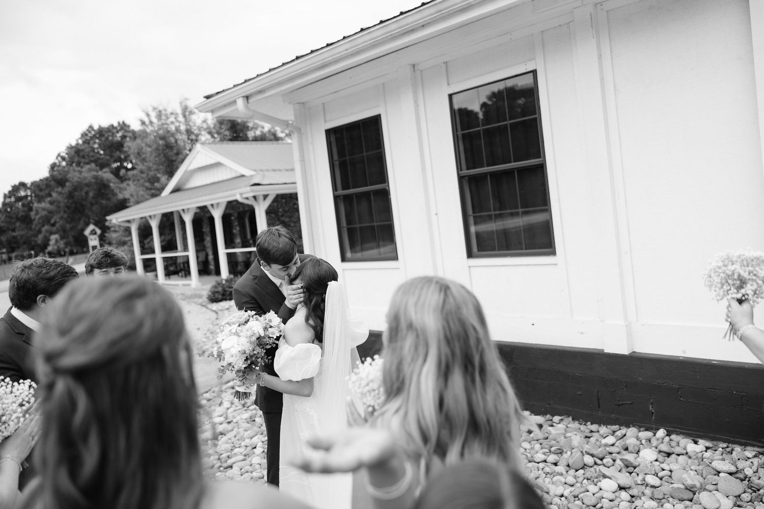 Black and white wedding photo of couple kissing outside a white building with guests watching.