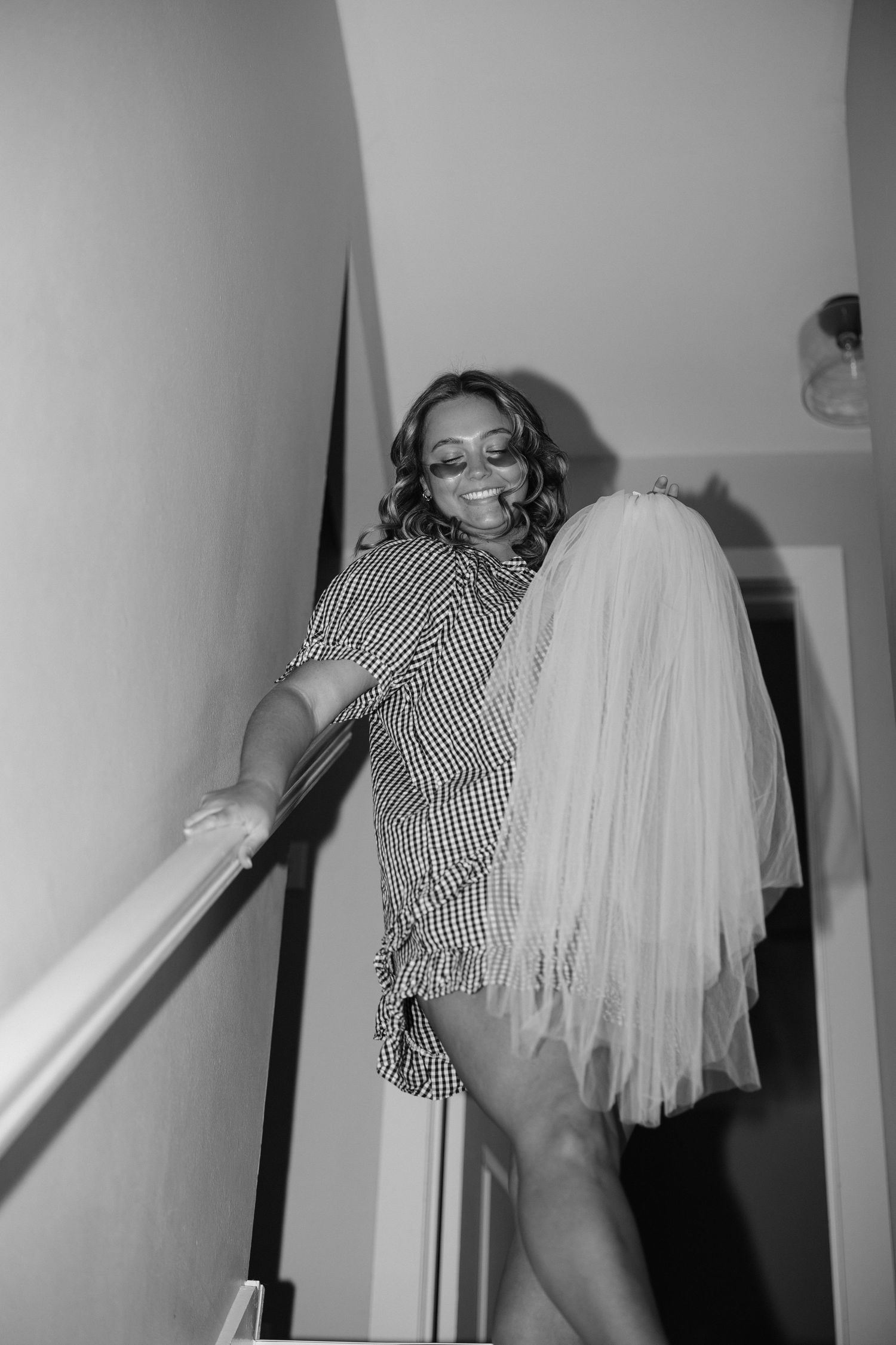 Black and white photo of a smiling bride holding a veil while descending stairs in a checkered dress and sunglasses.