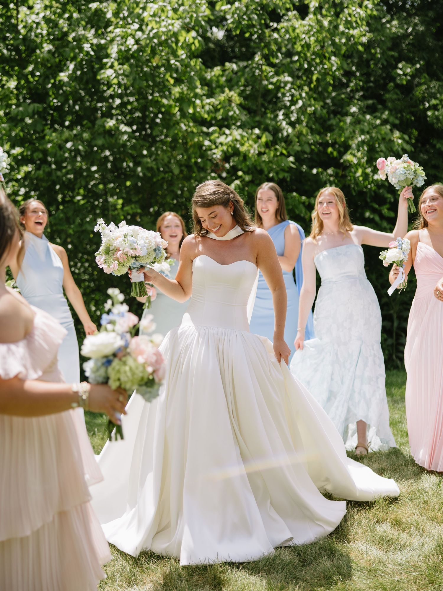 Bride in white ballgown twirls outdoors surrounded by bridesmaids in pastel dresses holding bouquets.