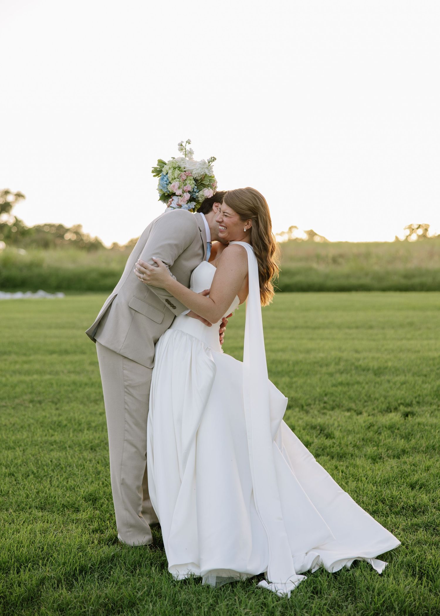 A couple shares a romantic kiss in a green field, bride in white gown holding floral bouquet, groom in tan suit.