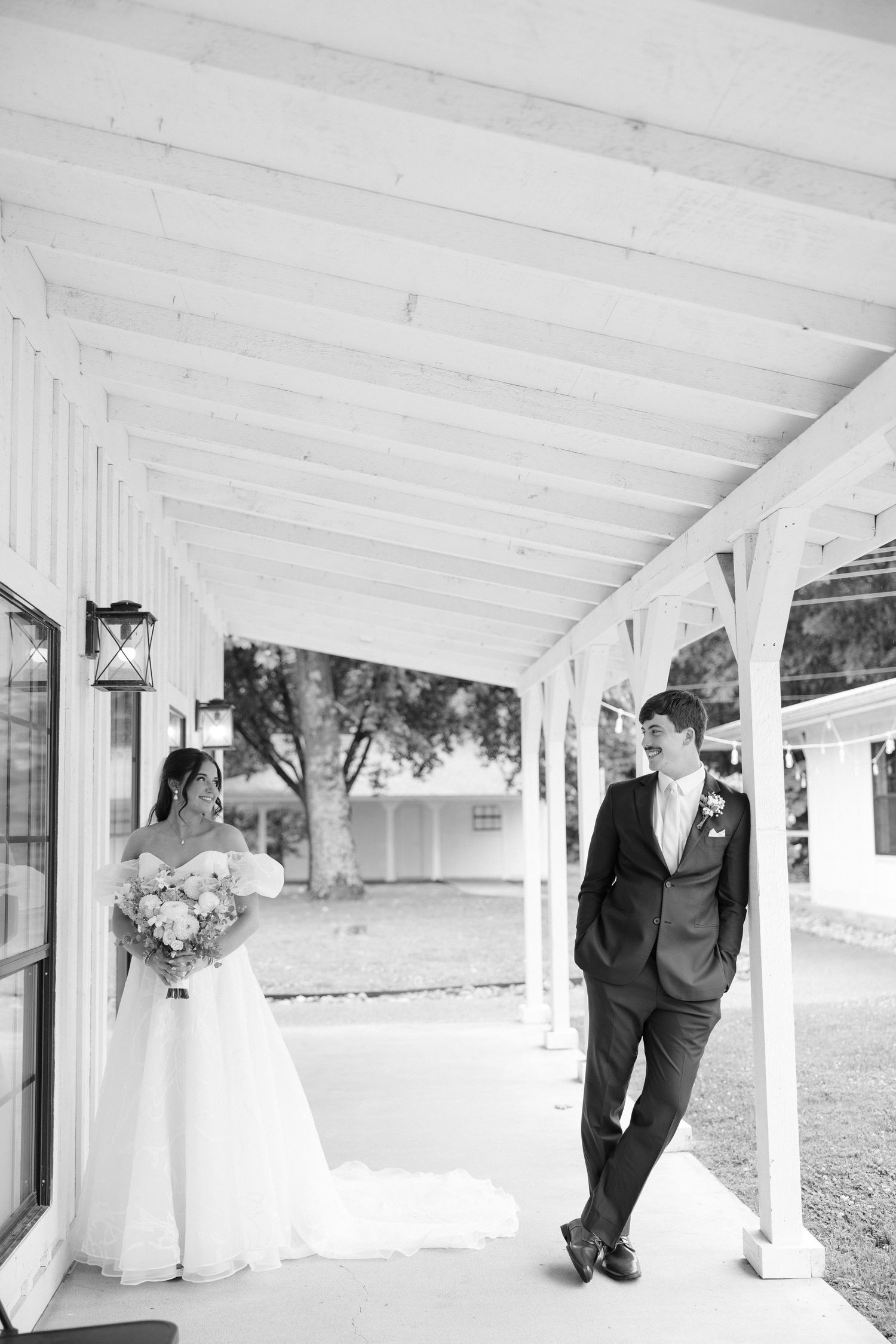 Black and white photo of a bride holding bouquet and groom in suit sharing a moment on a white covered porch.