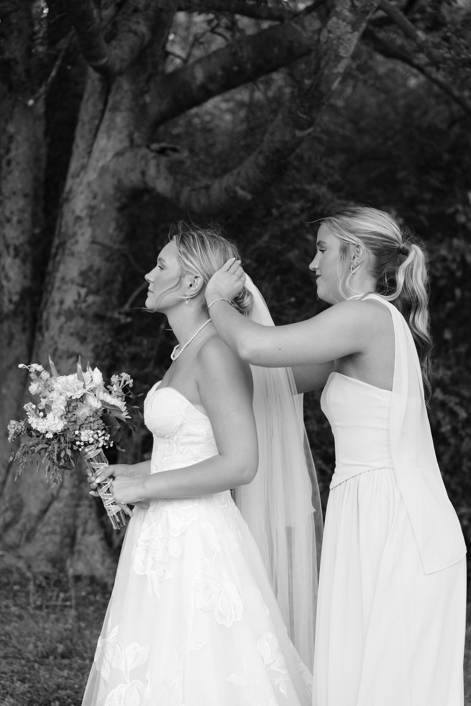 Bridesmaid adjusting bride's veil and necklace outdoors in black and white wedding photo.