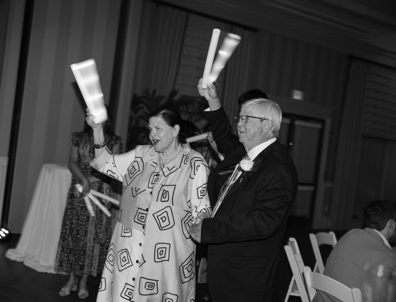 Elderly couple dancing joyfully at a formal event, raising glowing batons in a black and white photo.