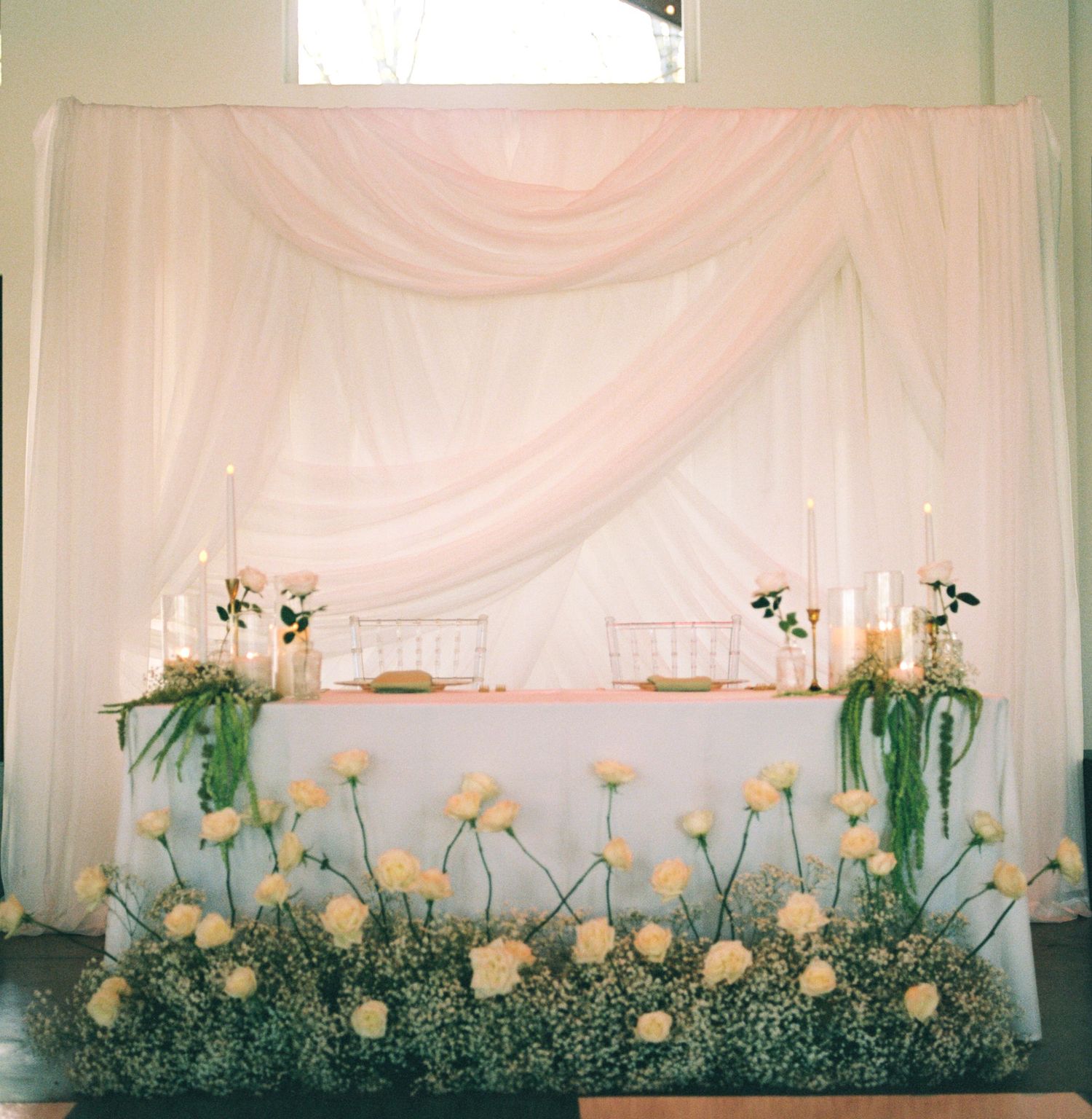 Romantic sweetheart table with blush draping, candles, and white rose floor arrangement.
