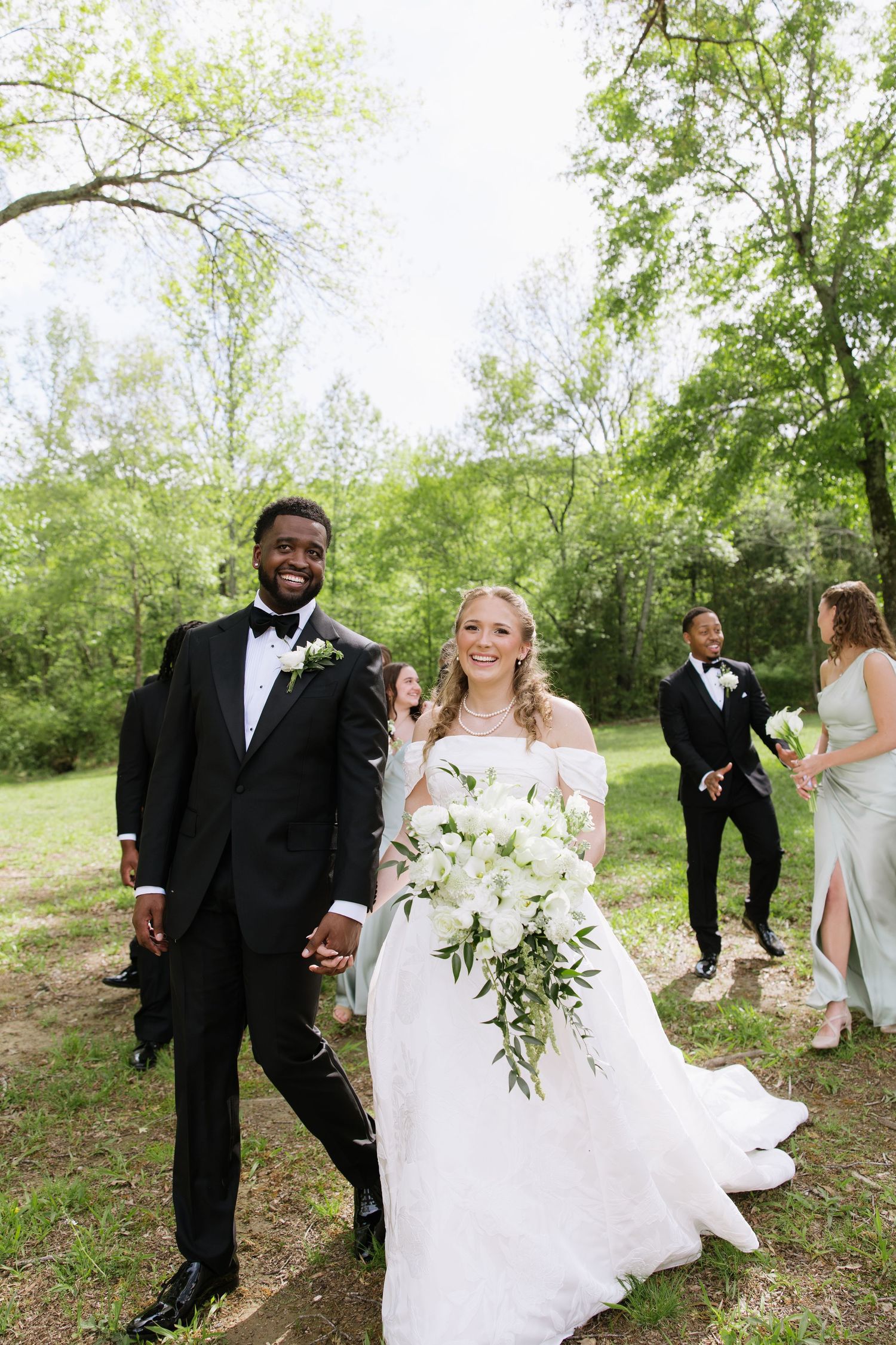 Interracial bride and groom walking hand-in-hand outdoors with wedding party, bride holding large white floral bouquet.