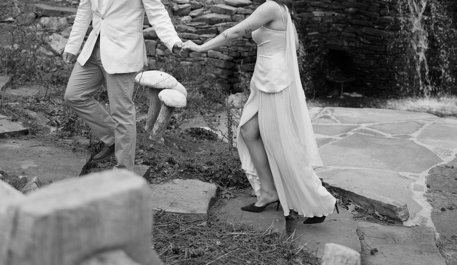 Black and white photo of couple holding hands while crossing stepping stones near a waterfall.