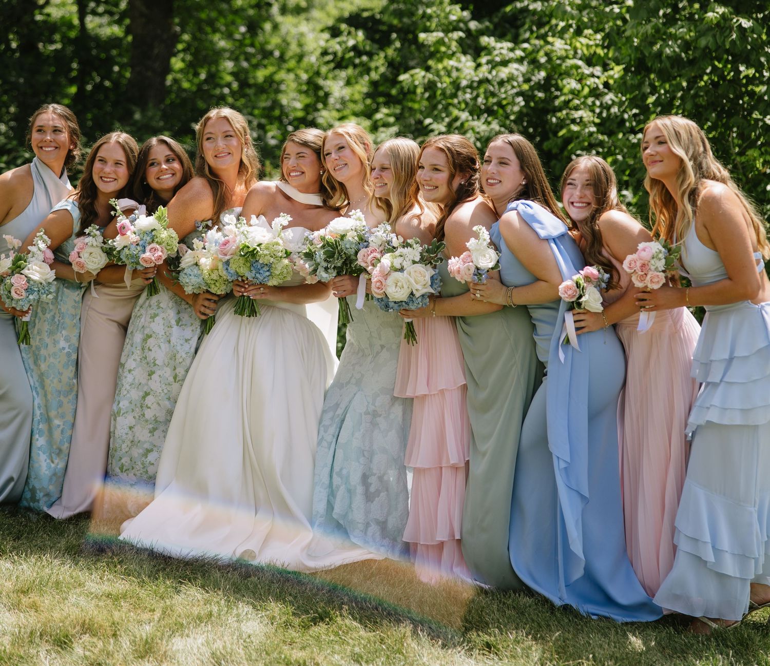 Bride and bridesmaids in pastel gowns holding floral bouquets, smiling outdoors on a sunny wedding day.
