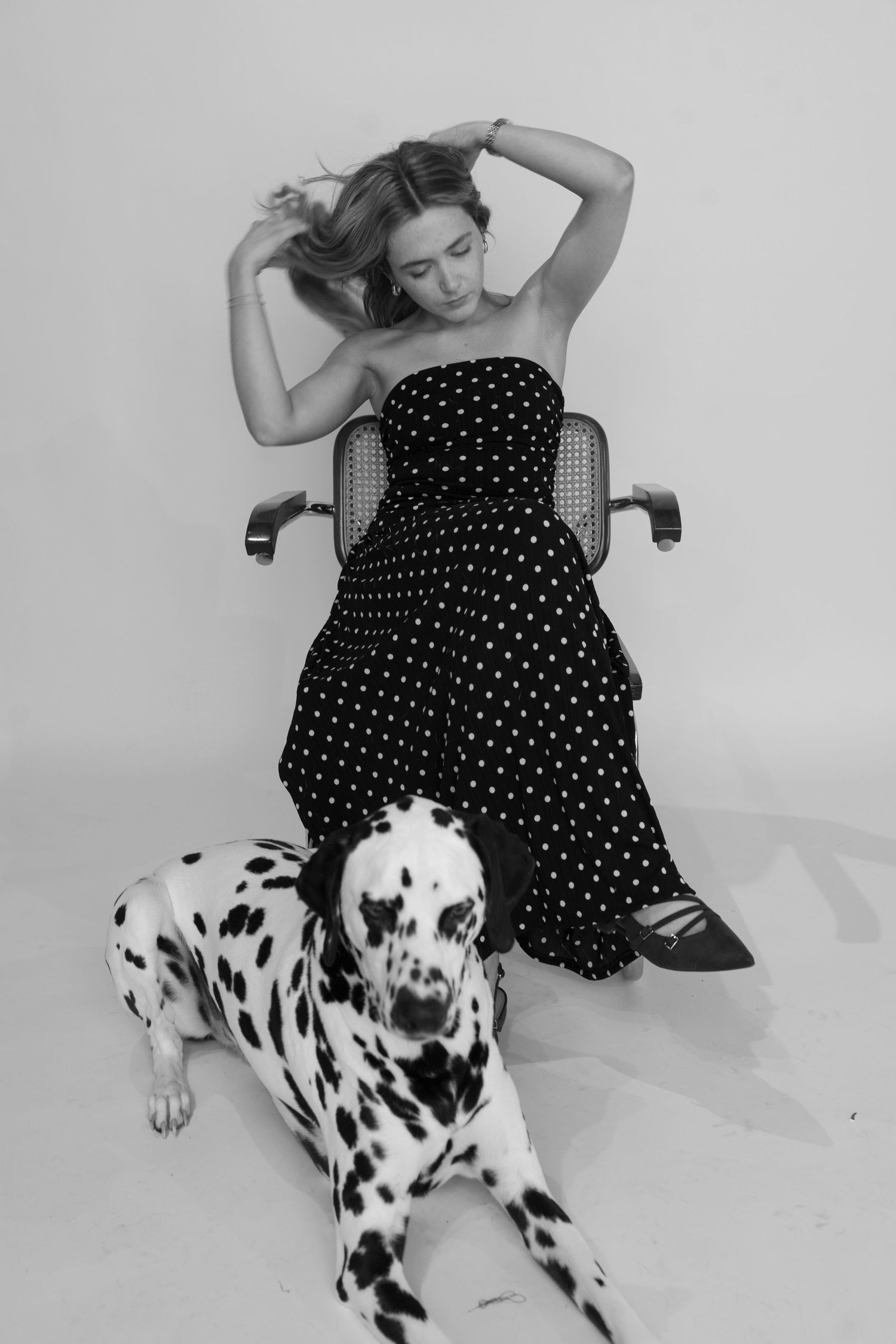 Woman in polka dot strapless dress sitting in chair with a Dalmatian dog lying at her feet, black and white photo.