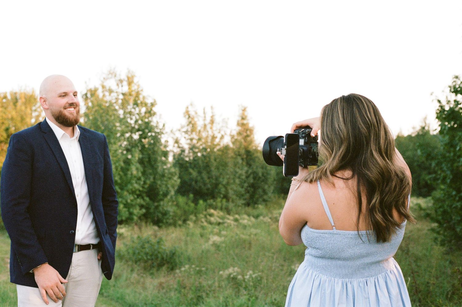 Woman in blue dress photographs smiling bald man in navy blazer outdoors in a sunny green field.