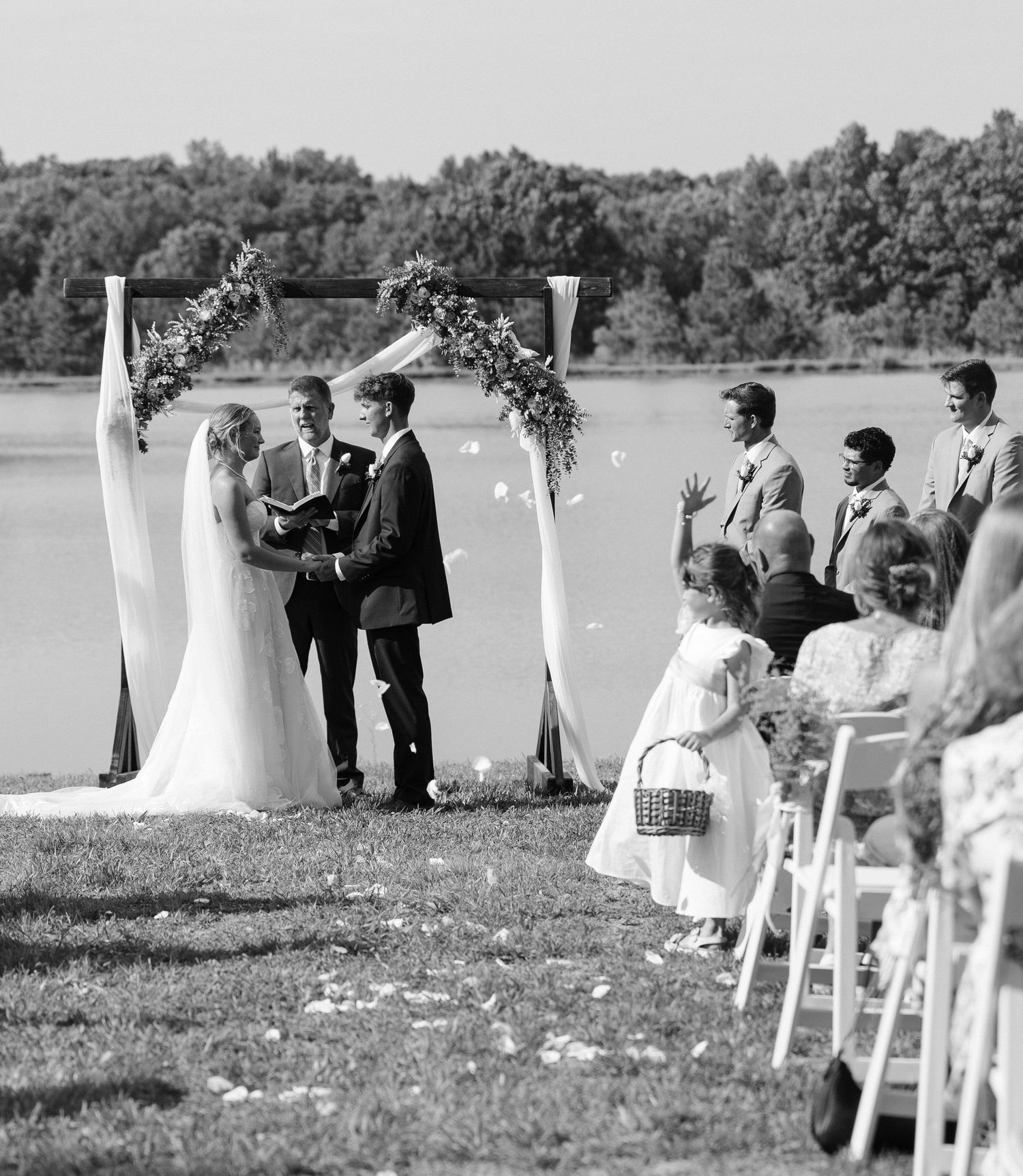 Black and white outdoor wedding ceremony by a lake with bride, groom, officiant, groomsmen, and flower girl.