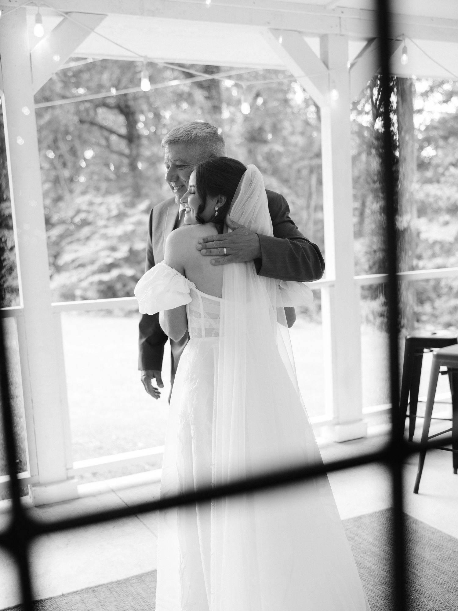 Black and white photo of a bride and her father sharing an emotional embrace under a gazebo on her wedding day.