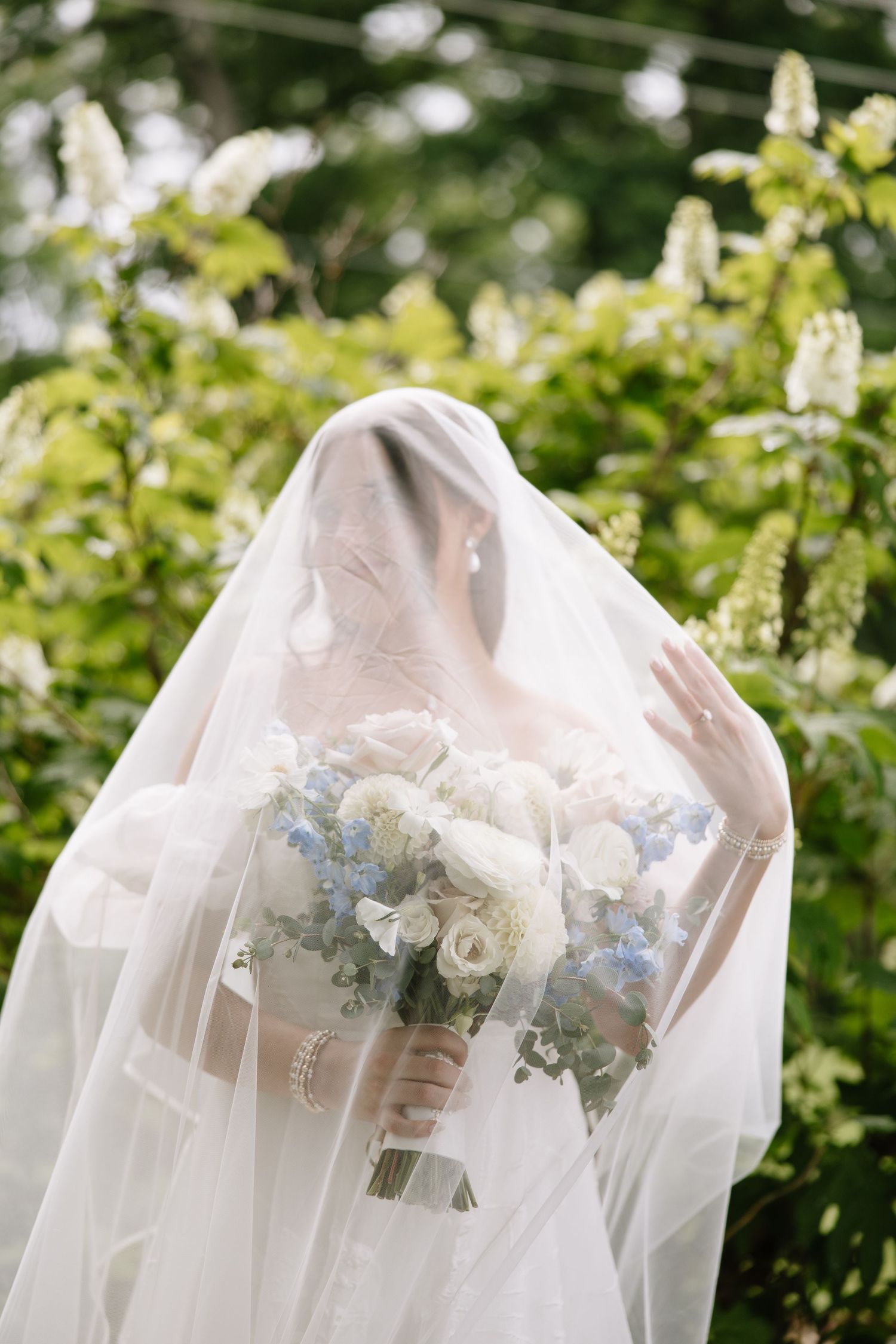 Bride holding a white and blue floral bouquet, face covered by a flowing veil, surrounded by lush green foliage.