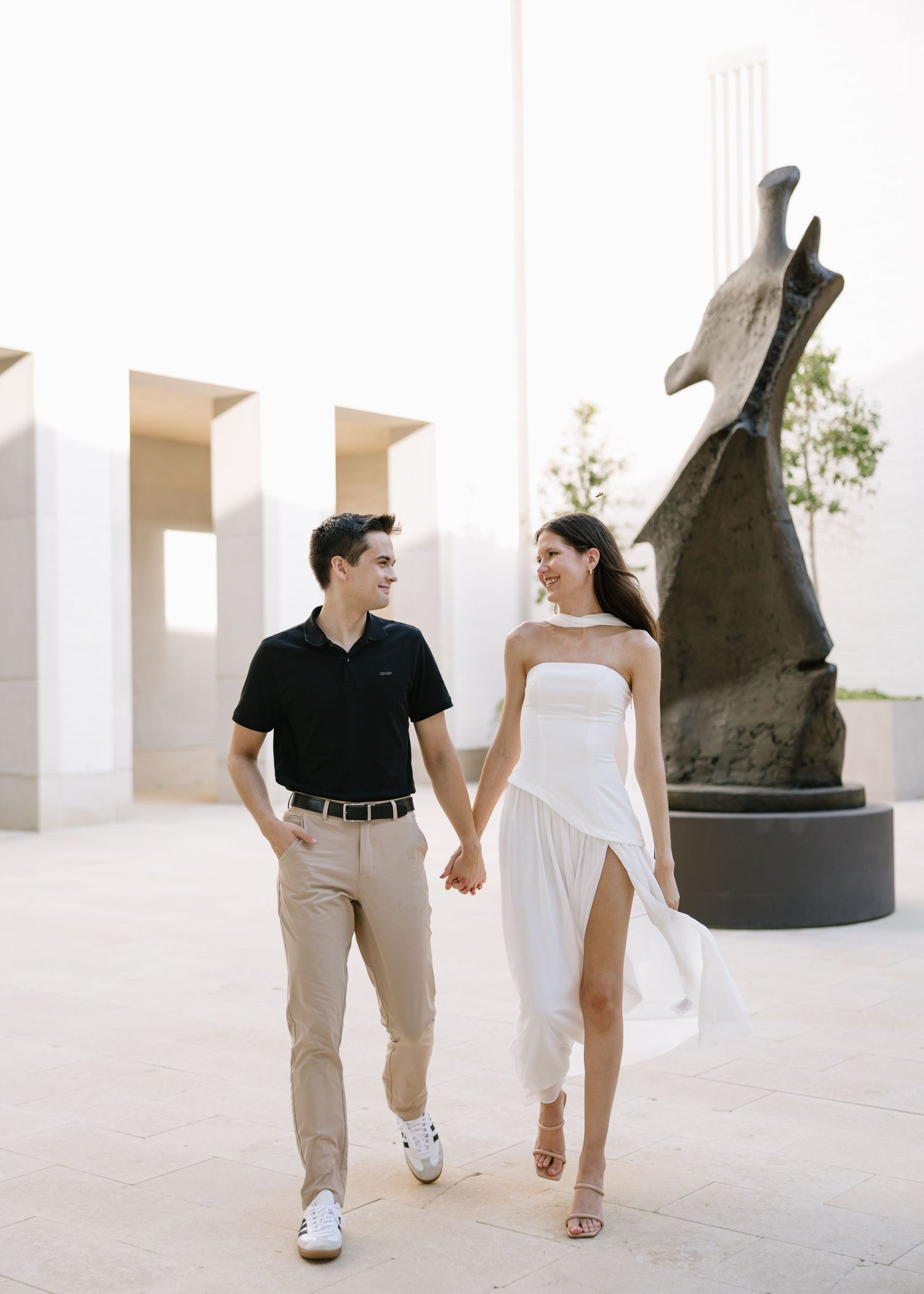 A couple holds hands and smiles while walking outdoors near a modern sculpture in elegant attire.