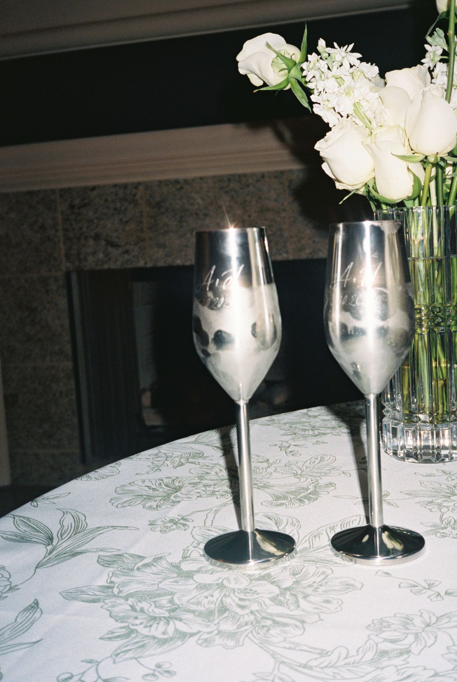 Two silver champagne flutes on a floral tablecloth beside a vase of white roses, perfect for a wedding toast.