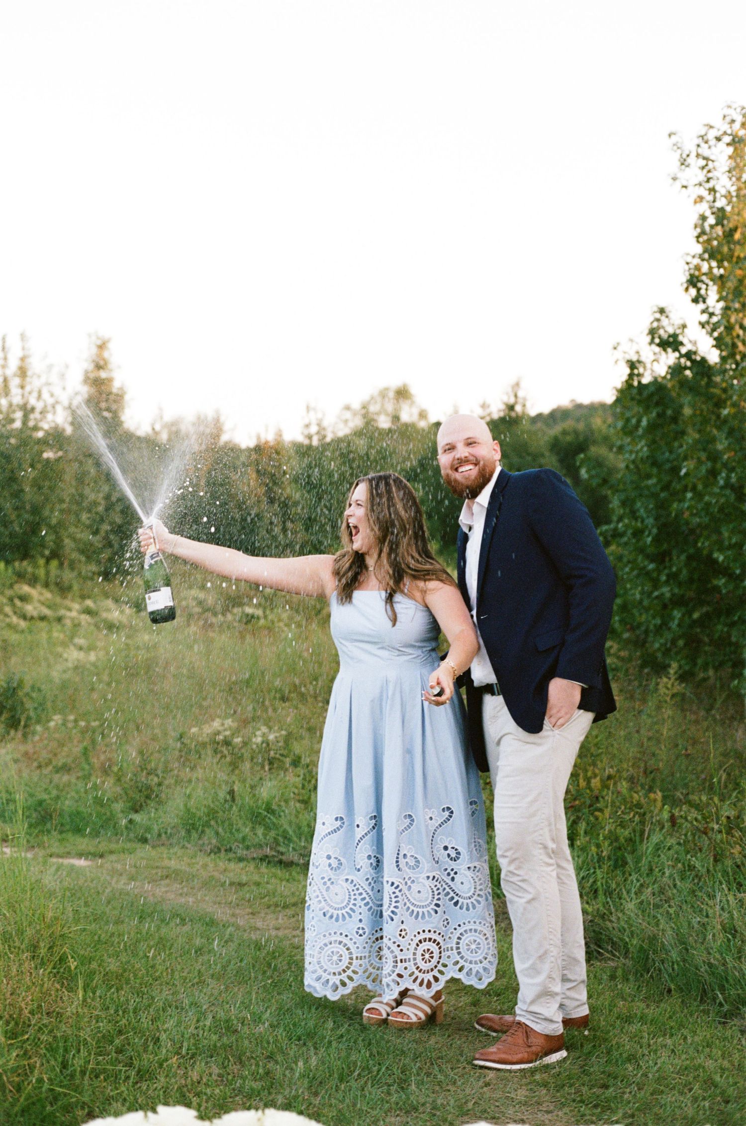 A joyful couple celebrates outdoors, woman in blue dress spraying champagne while man in navy blazer embraces her in a green field.