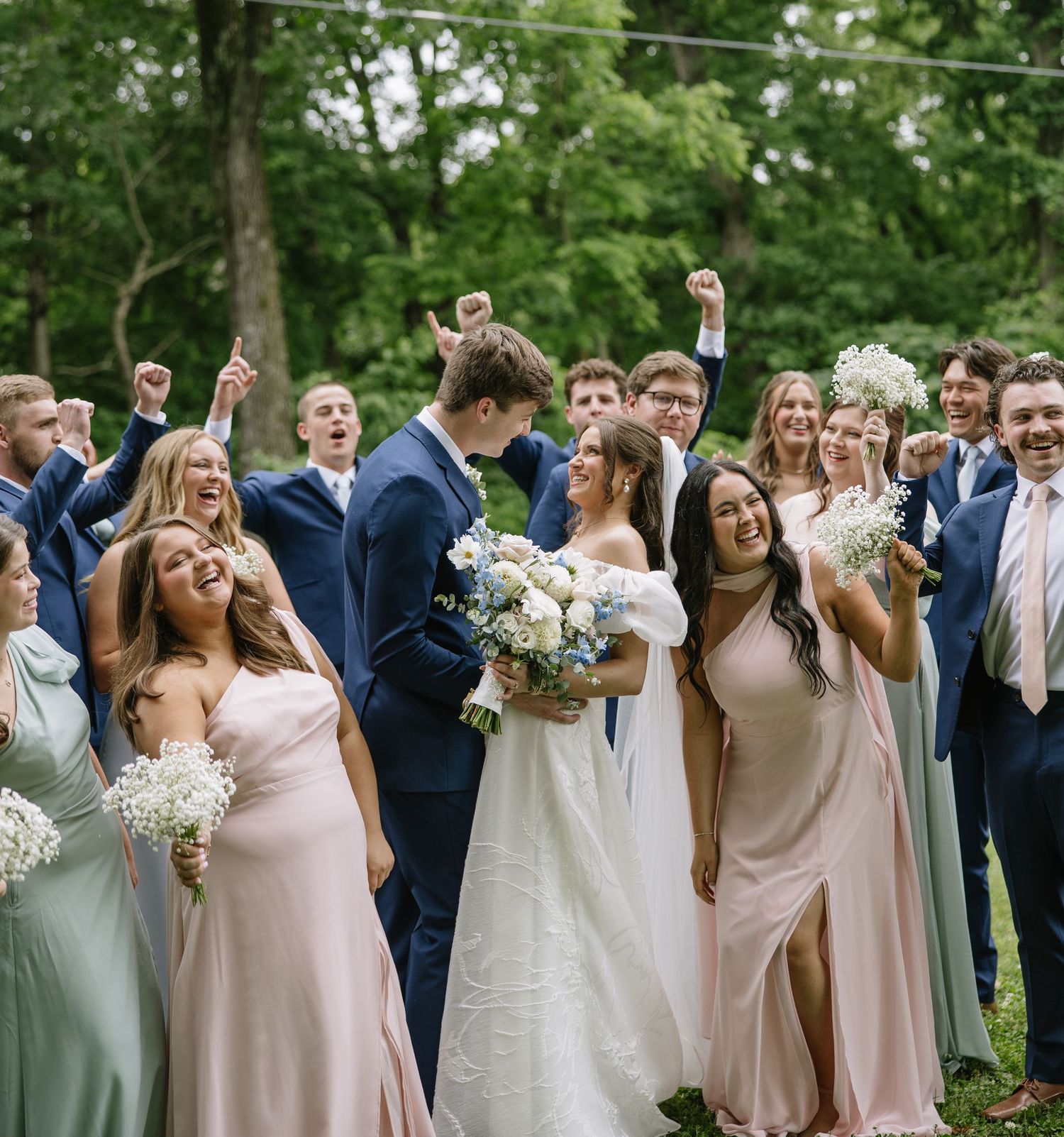 Joyful wedding party celebrating outdoors with bride in white gown holding bouquet, bridesmaids in pink and sage, groomsmen in navy suits.