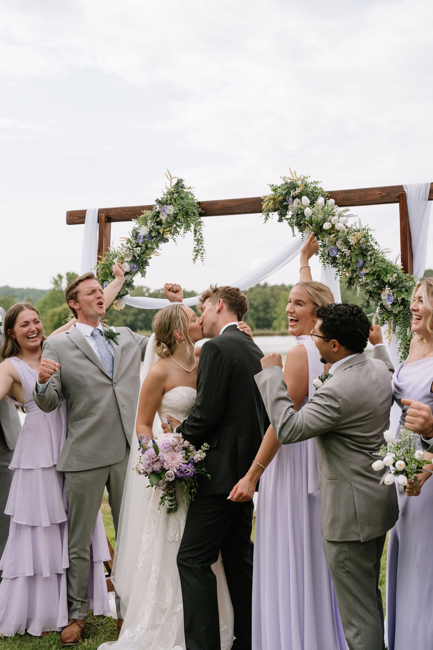 Bride and groom share first kiss under floral arch surrounded by cheering wedding party in lavender dresses and gray suits.