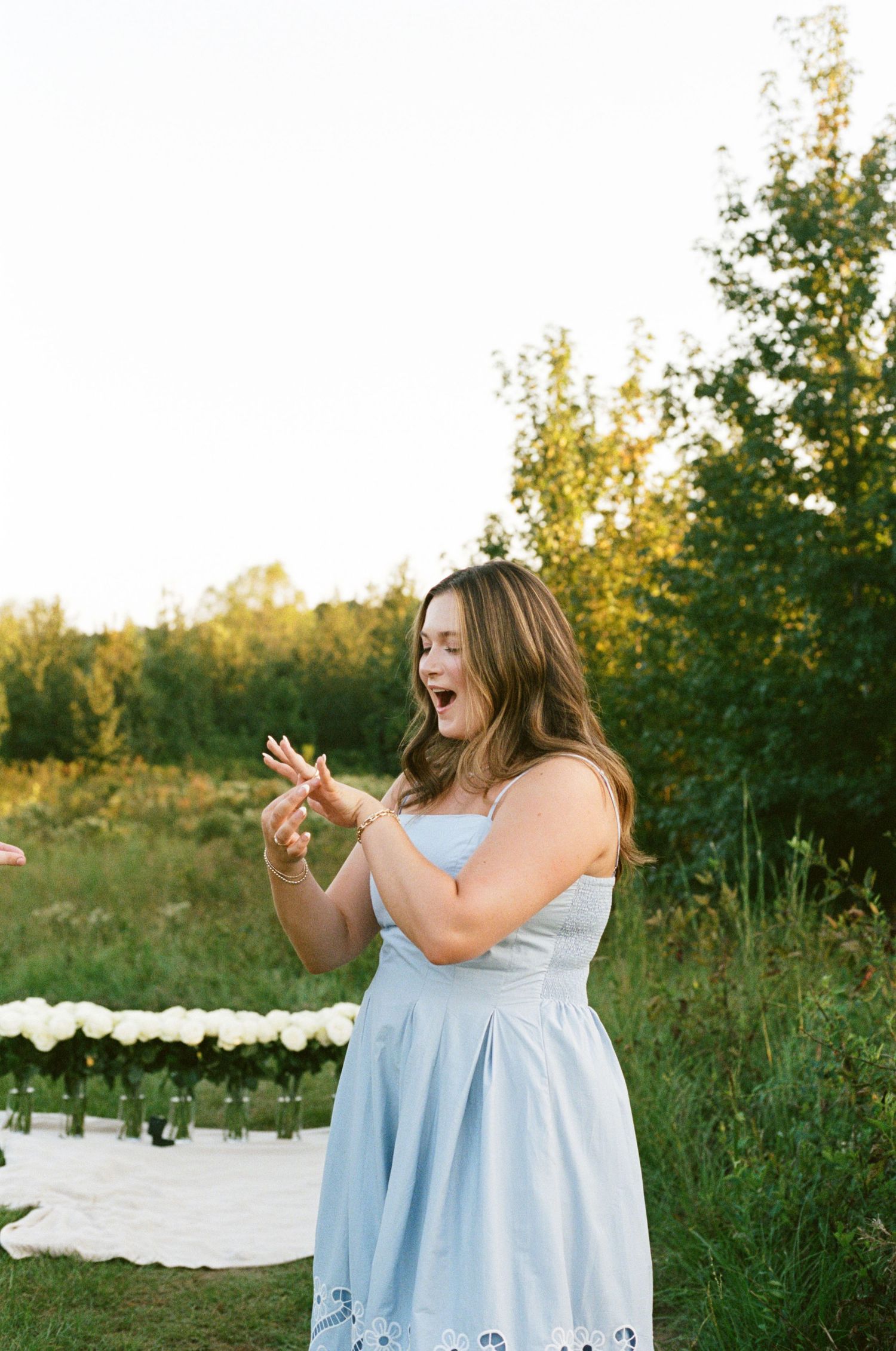 Woman in light blue sundress clapping outdoors at golden hour with white floral arrangement and greenery in background.
