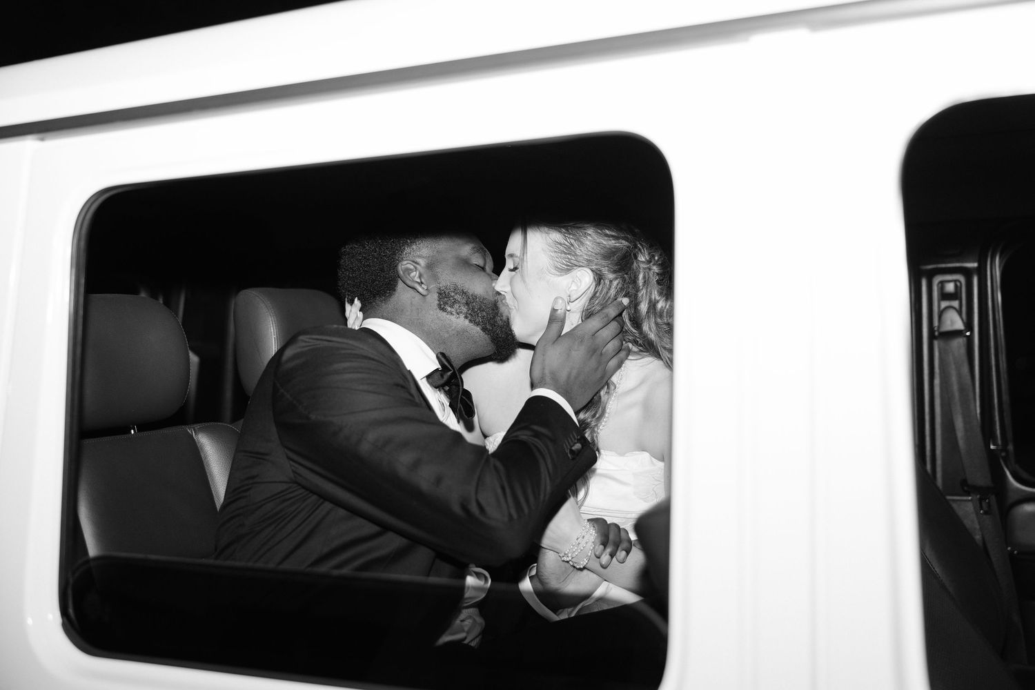 Black and white intimate photo of newlyweds sharing a kiss inside a white limousine after their wedding.