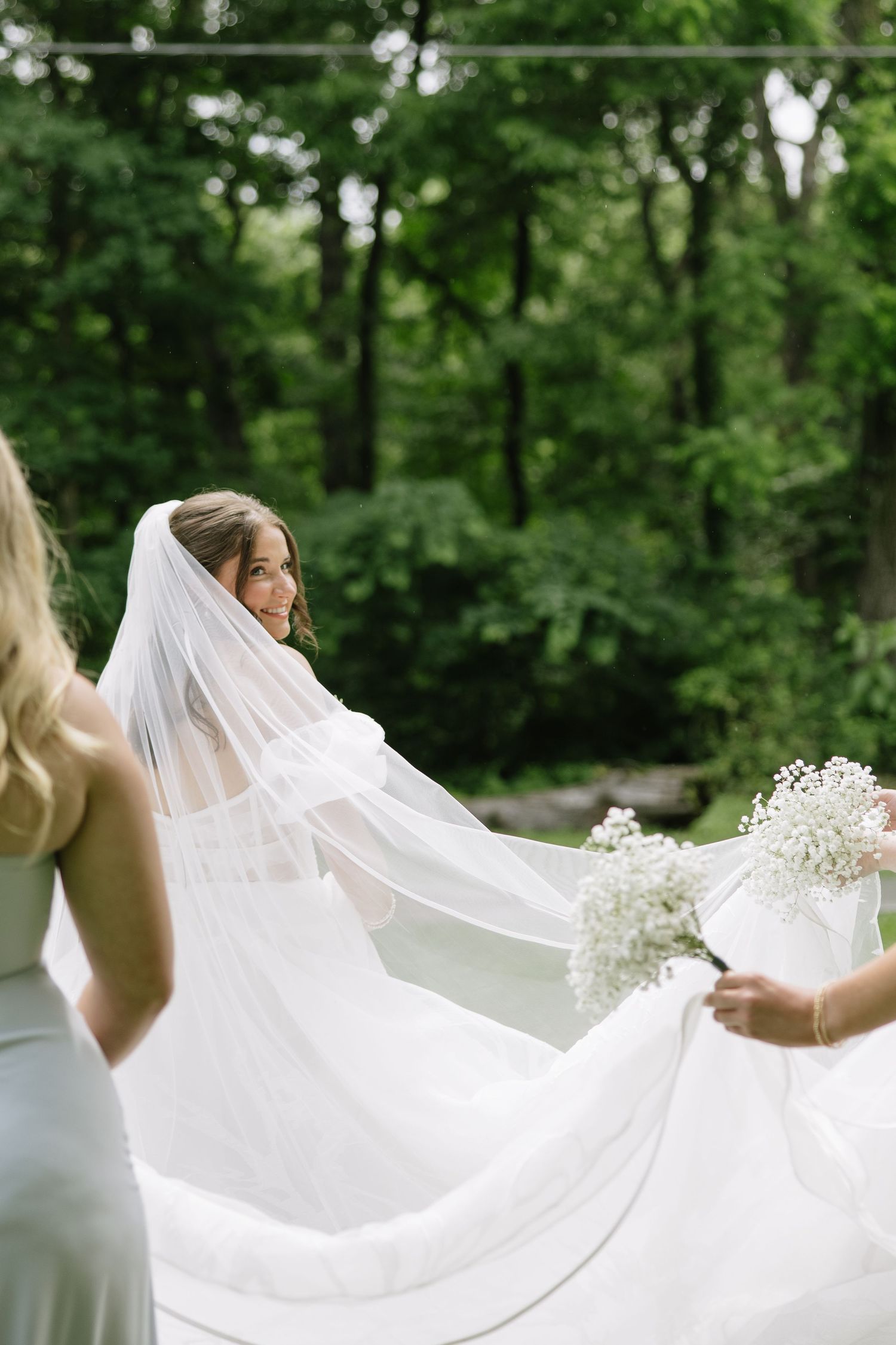 Bride in white gown and veil smiles back at bridesmaids holding her train outdoors among lush green trees.