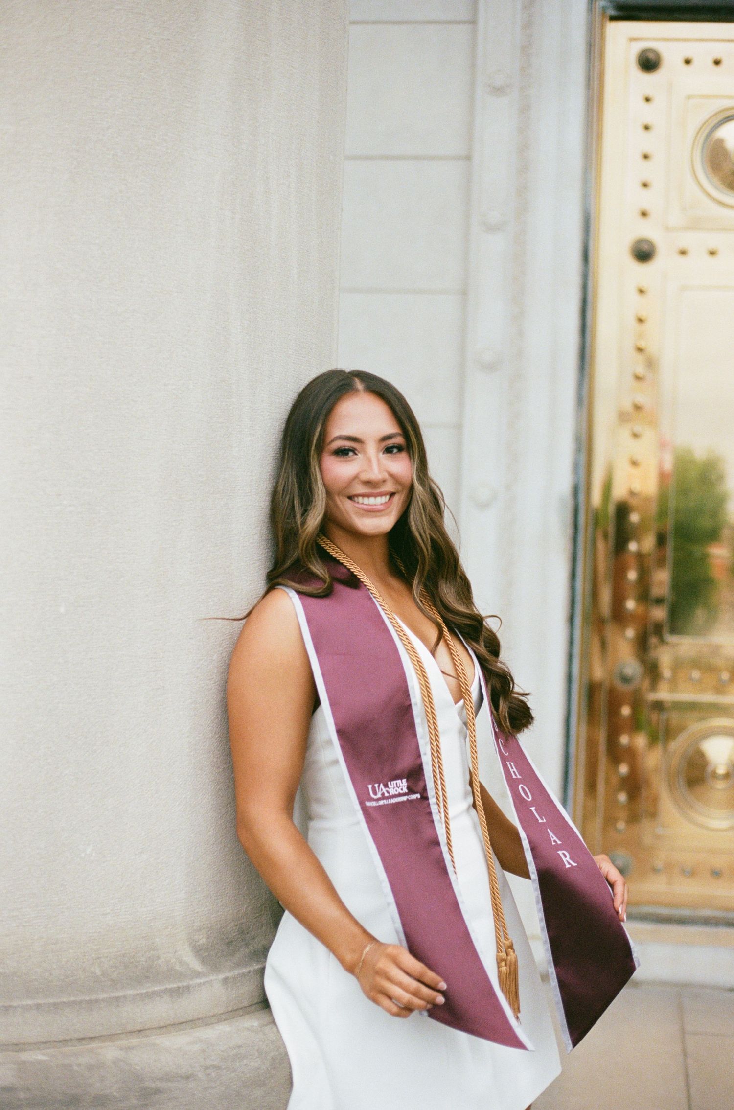 Smiling graduate wearing maroon honor sash poses against ornate column at graduation ceremony.