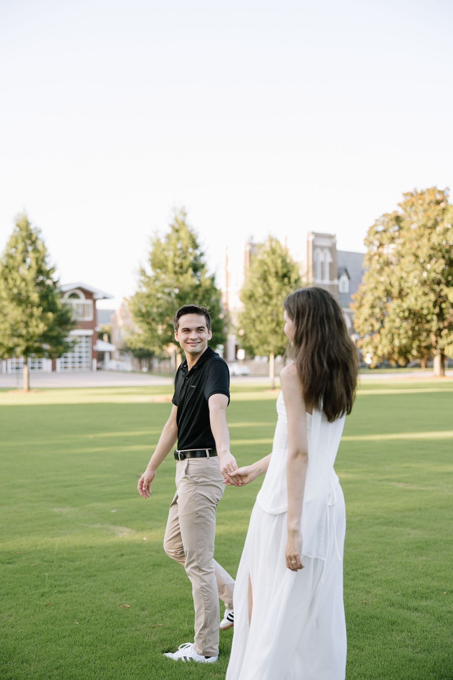 A smiling couple holds hands while walking on a green lawn, the man in black polo and khakis, woman in a white dress.