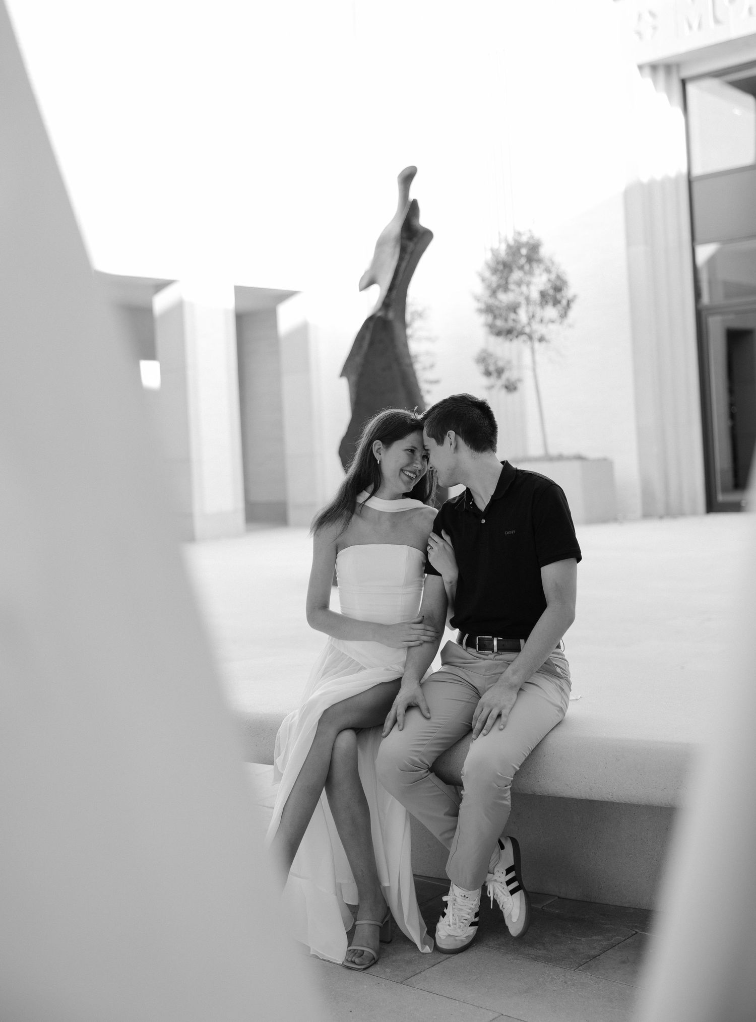 Black and white photo of a romantic couple sitting close together on a bench near a sculpture outdoors.
