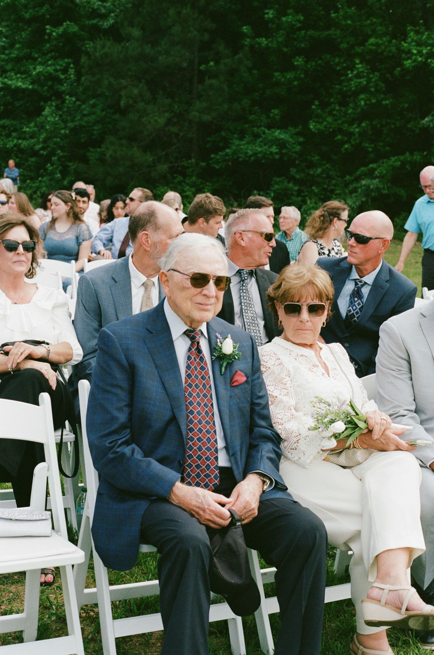 Elderly couple in formal attire seated at an outdoor wedding ceremony, man wearing blue suit with boutonniere and red tie.