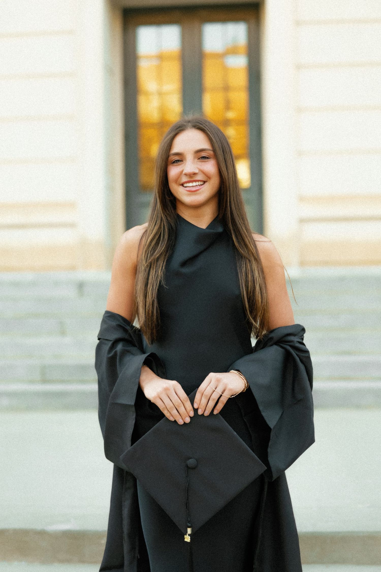 Happy graduate in black dress holding mortarboard cap while wearing graduation gown on university steps.