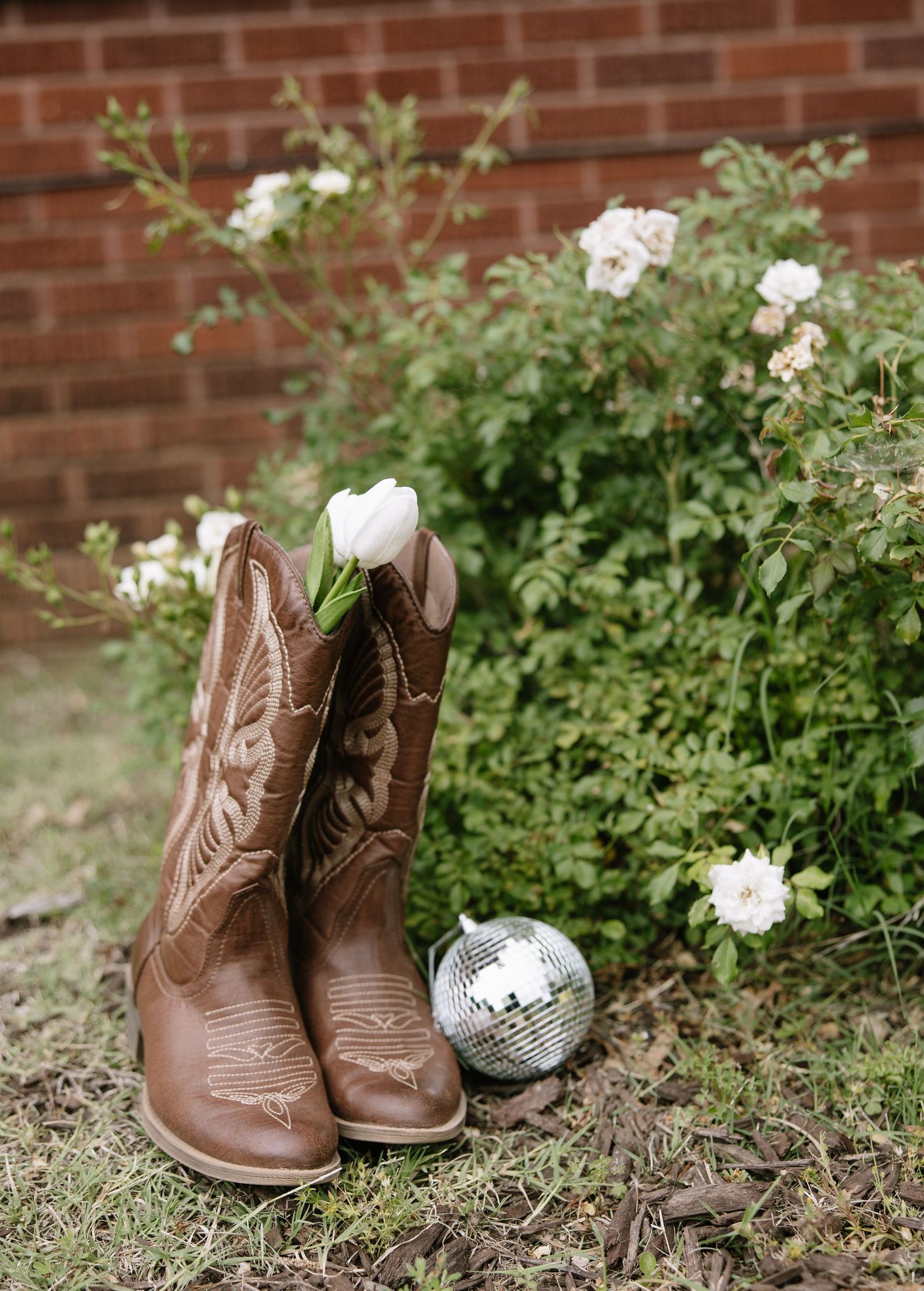 Brown cowboy boots with a white rose boutonniere and mini disco ball beside blooming white roses outdoors.