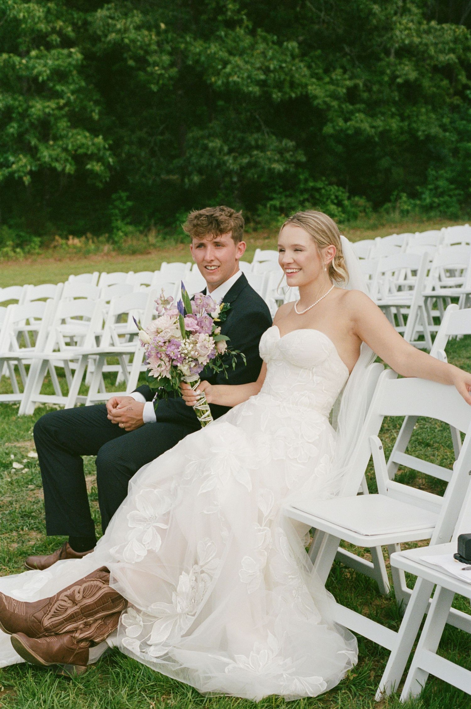 Bride in strapless gown with cowboy boots and groom in black suit sit together at outdoor wedding ceremony.