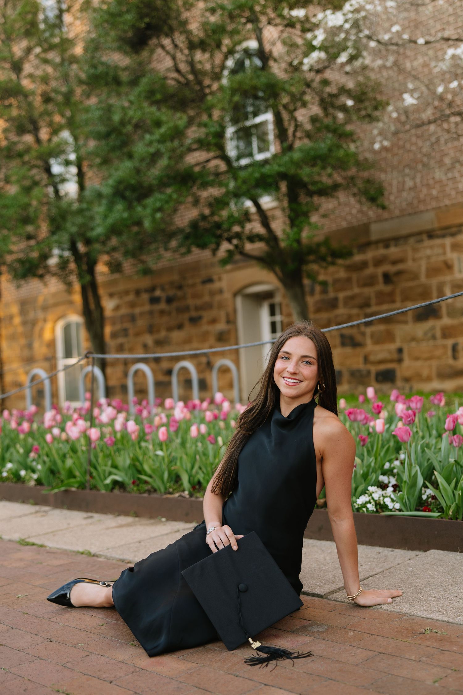 Smiling graduate in black dress sitting on brick steps with pink tulips and stone building in background.