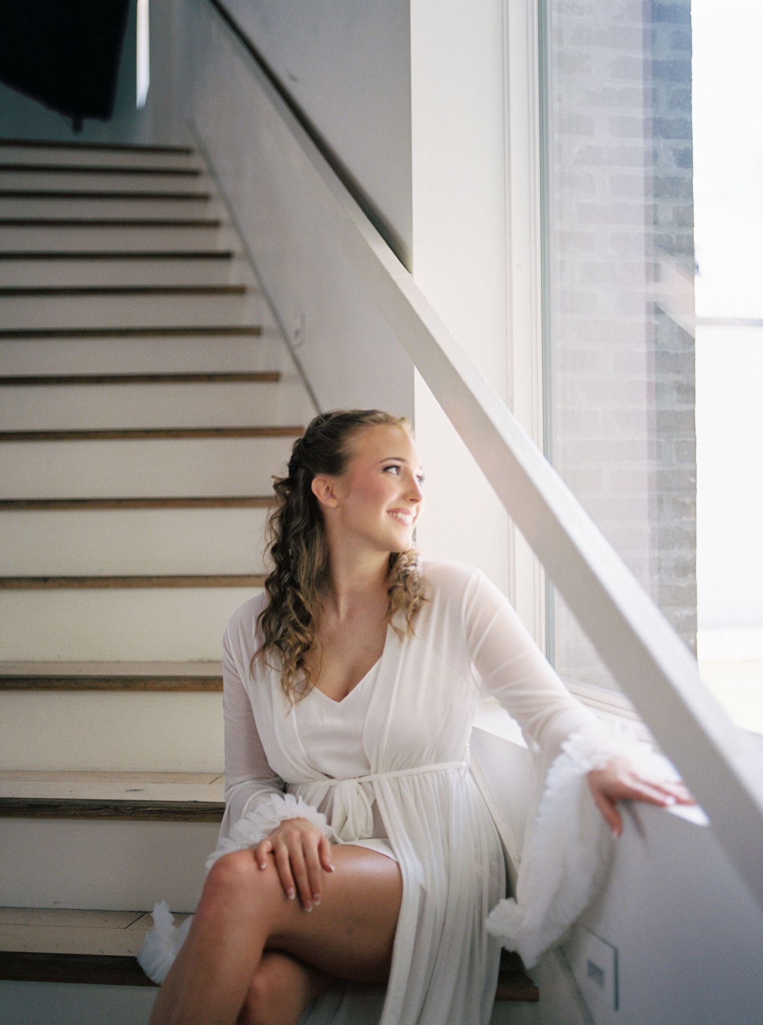 Smiling bride in white robe sitting on staircase by large window in soft natural light.